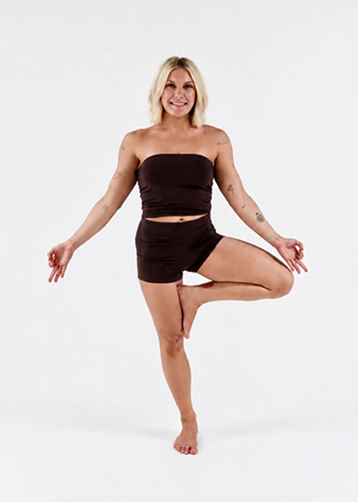 A woman with blonde hair in a yoga pose, balancing on one foot with her knee bent and her foot resting on her inner thigh, while holding her other leg parallel to the ground. She is smiling and wearing a dark strapless top and matching shorts, set against a plain white background.
