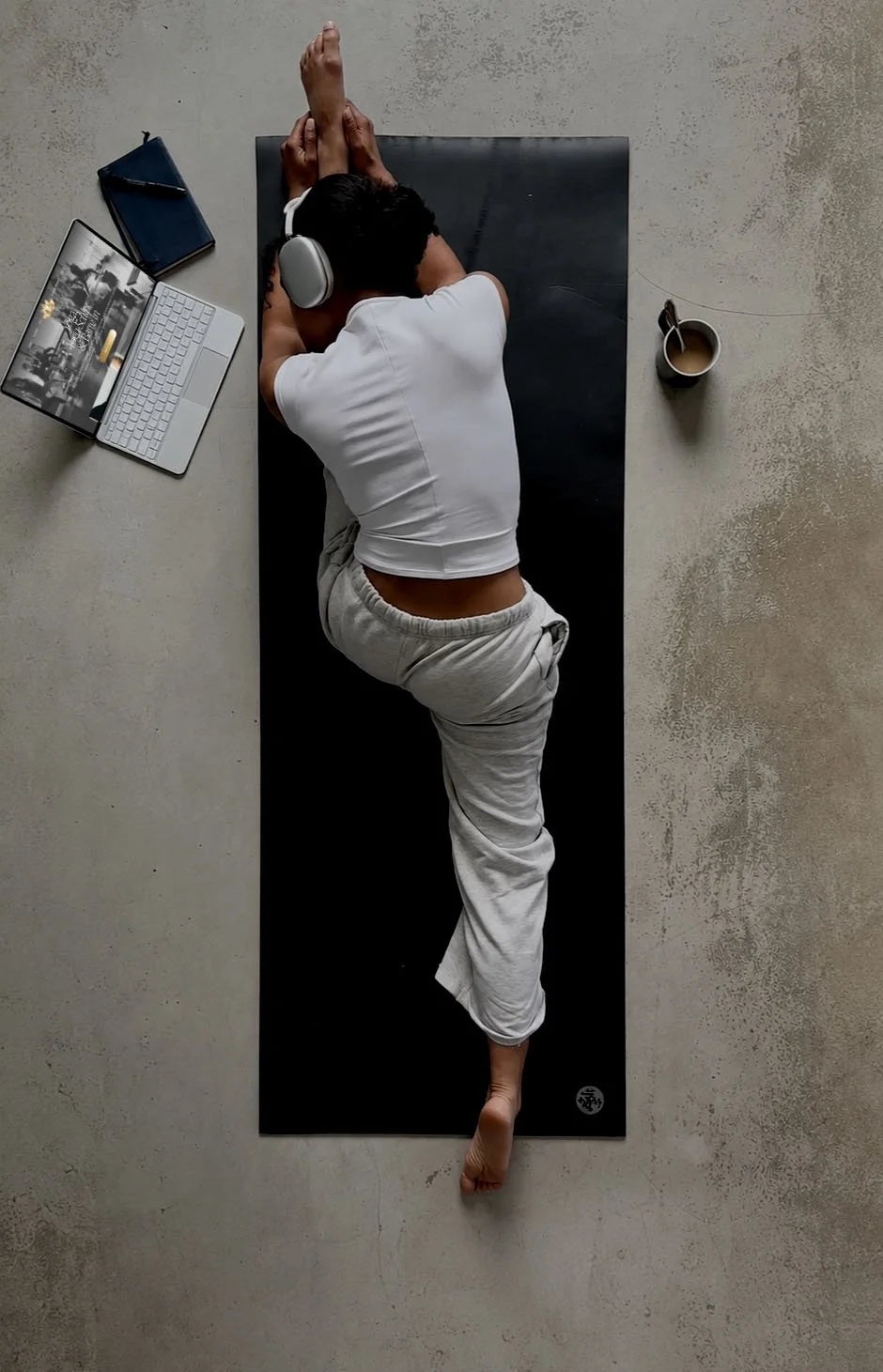 A person lying on their side on a black yoga mat, wearing a white t-shirt and light-colored sweatpants, with headphones on, resting at home with a laptop, a notebook, a coffee mug, and a spoon nearby.