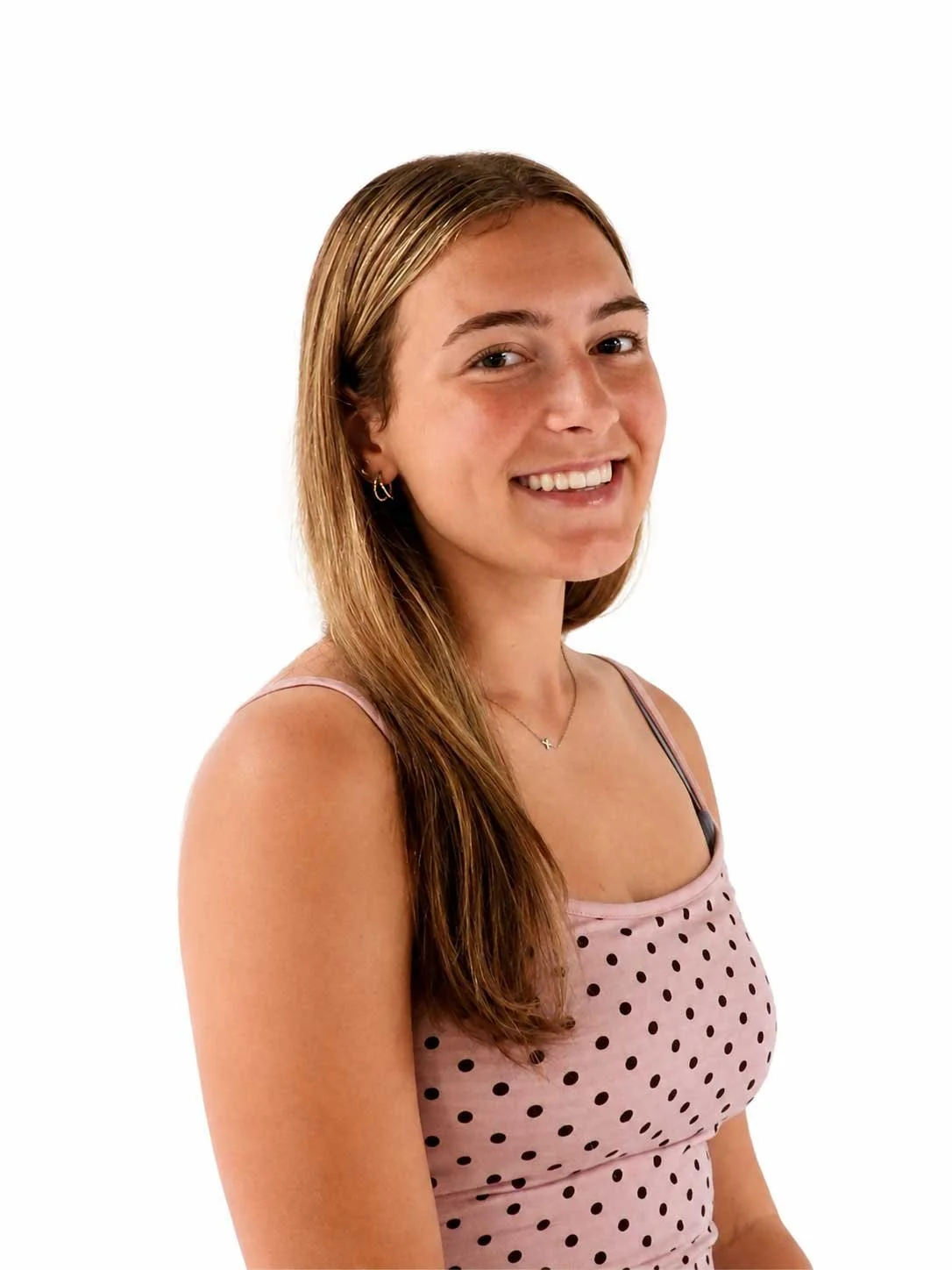 A young woman with long, light brown hair smiling, wearing a pink polka dot tank top, a necklace, and small hoop earrings, against a plain white background.