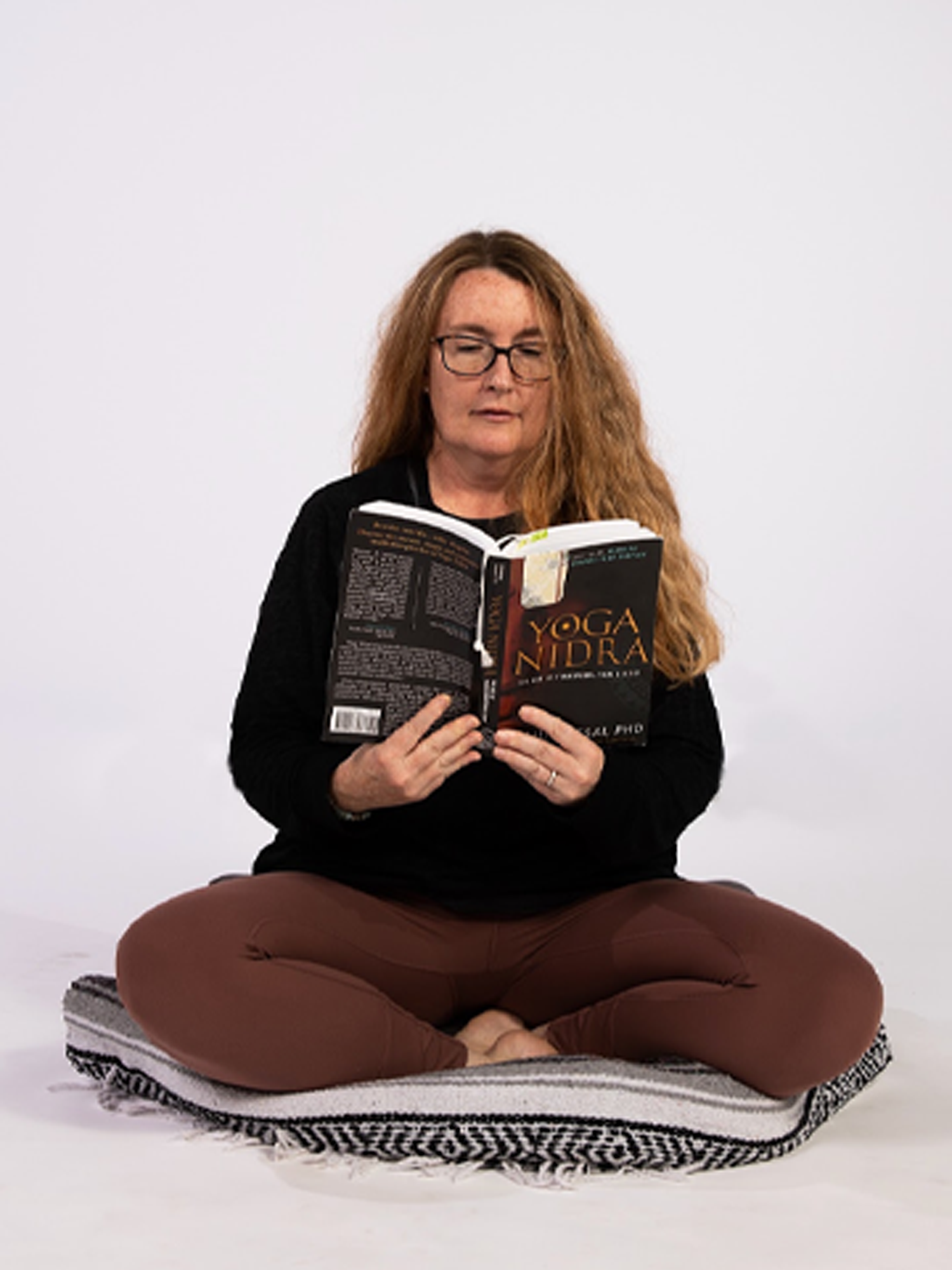 A woman with long red hair, glasses, and light skin sitting cross-legged on a blanket, reading a book titled 'Yoga Nidra' in front of a plain white background.