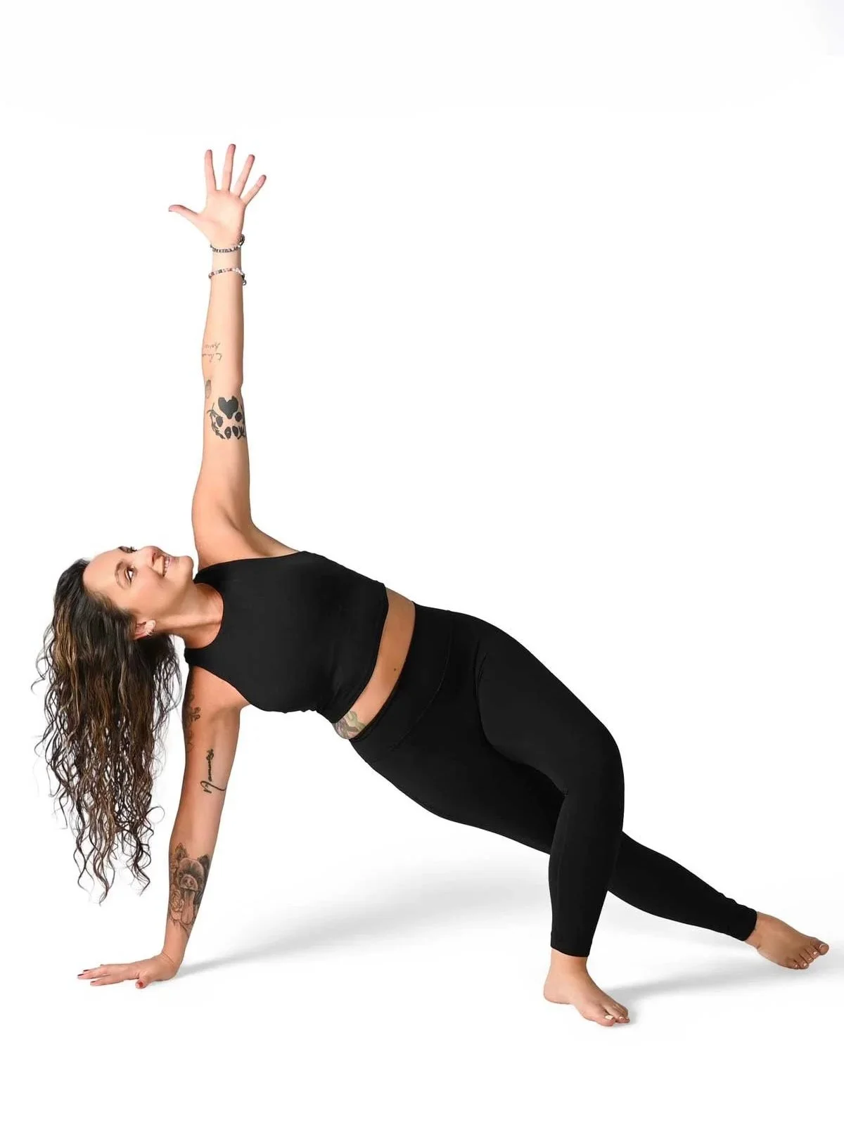 A woman with curly hair doing a side plank yoga pose on a white background, wearing a black sports bra and black leggings.