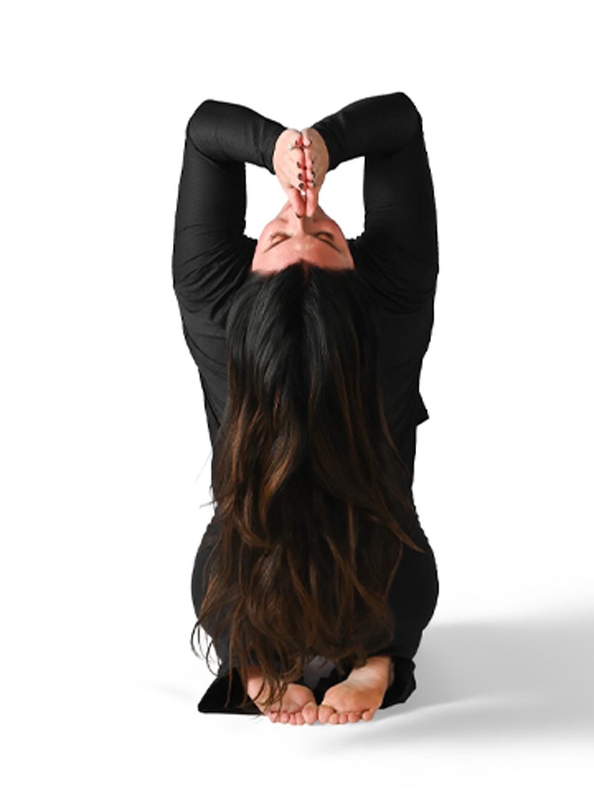 Woman in black long-sleeve top performing a yoga pose, kneeling on the ground with arms raised above head, hands pressed together in prayer position, with long dark hair hanging down.