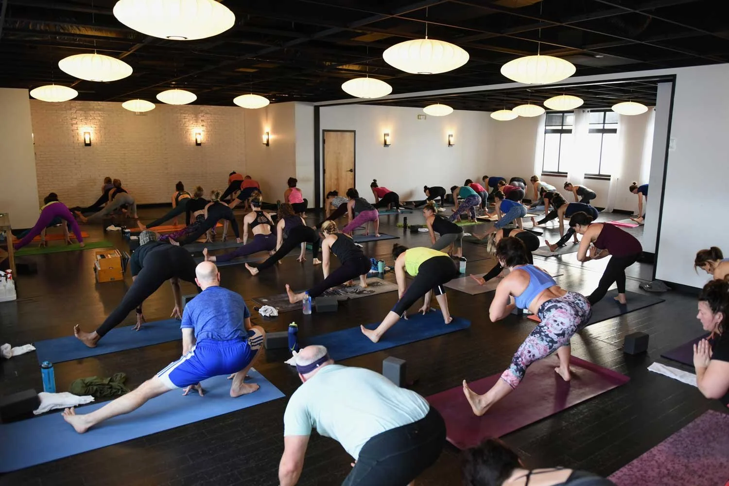 A large group of people participating in a yoga class in a spacious, well-lit studio with black hardwood floors, white brick walls, and round ceiling lights. They are engaged in various yoga poses on mats, with some people using yoga blocks and water bottles nearby.