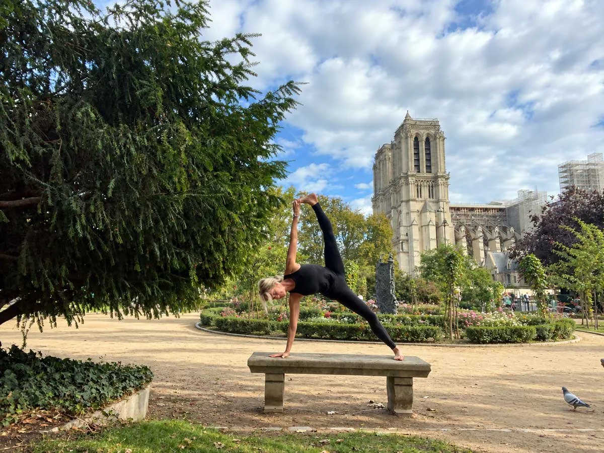 A woman performing a yoga pose on a wooden bench in a park with trees, flowers, and the Notre-Dame Cathedral in the background on a partly cloudy day.