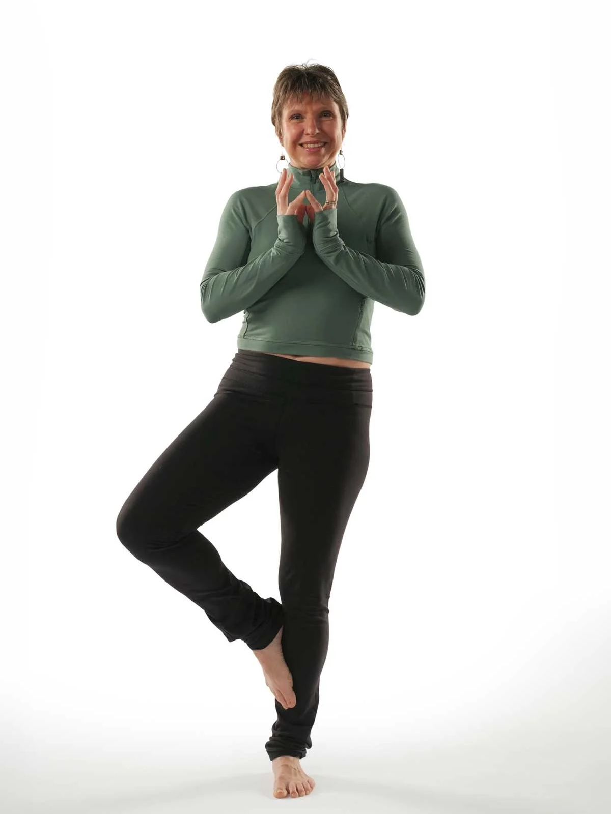 A woman standing on one leg, with arms raised and hands clasped in a yoga pose, wearing a green top and black leggings, smiling against a white background.