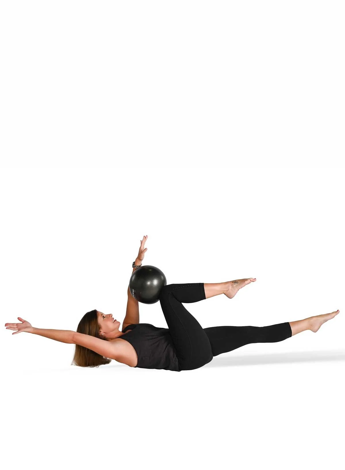 A woman performing an exercise on the floor with a black fitness ball, lying on her back with her left arm extended overhead and her right arm holding the ball near her bent right knee, which is raised. Her left leg is extended straight on the ground.