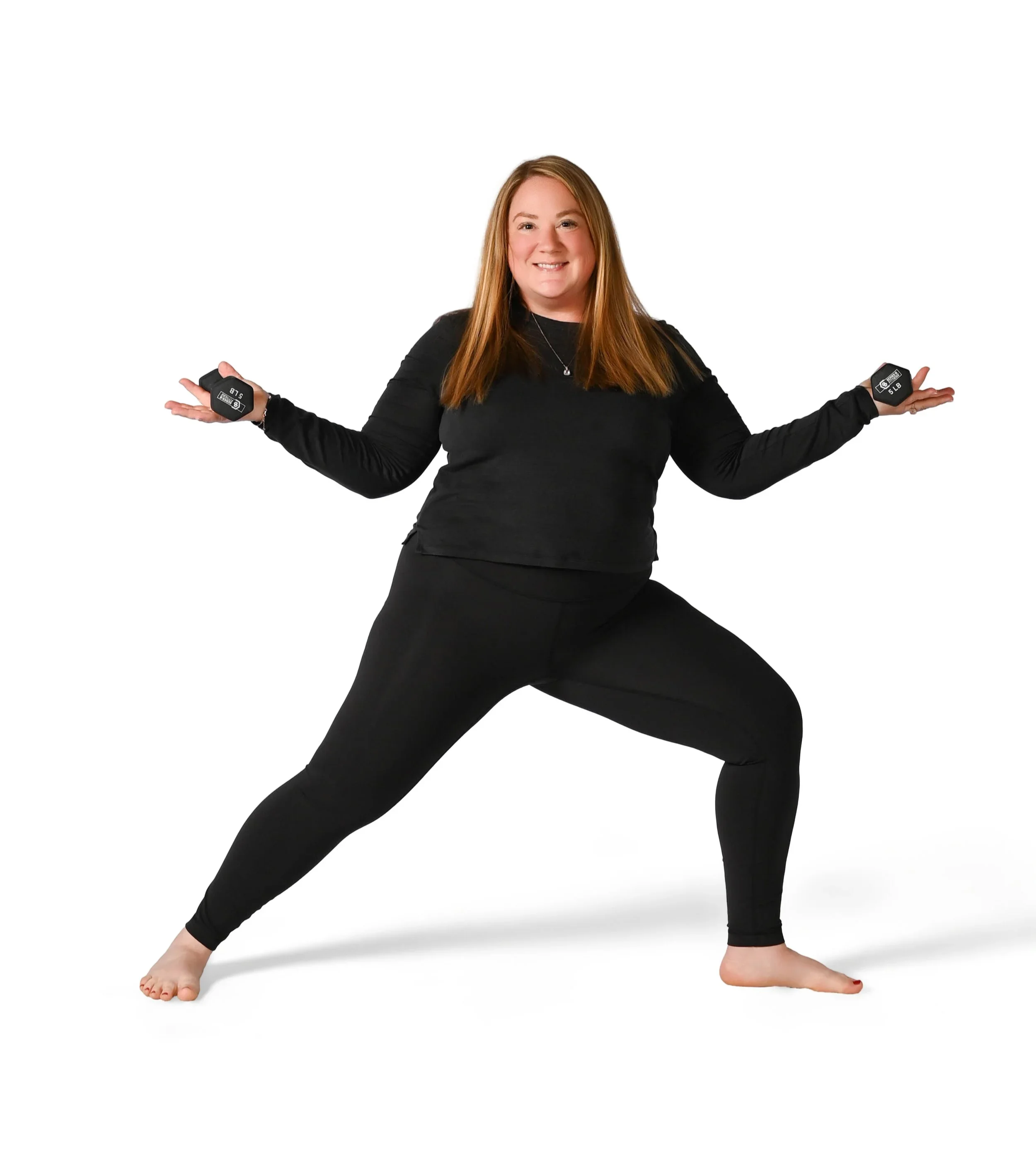 Woman in black long sleeve top and black leggings practicing yoga in warrior pose with arms outstretched and mouth smiling, against a plain white background.