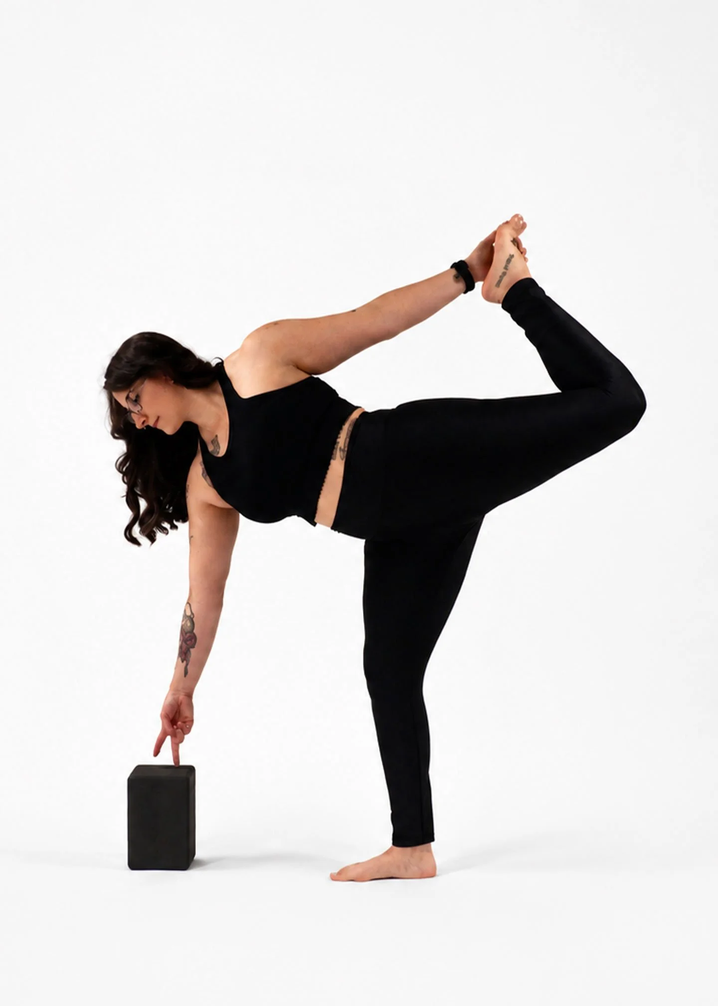 A woman in black leggings and a black tank top balances on one leg in a yoga pose, touching a black block with one hand and holding her foot with the other hand behind her back. She has long brown hair, glasses, and tattoos on her arm and shoulder, against a plain white background.