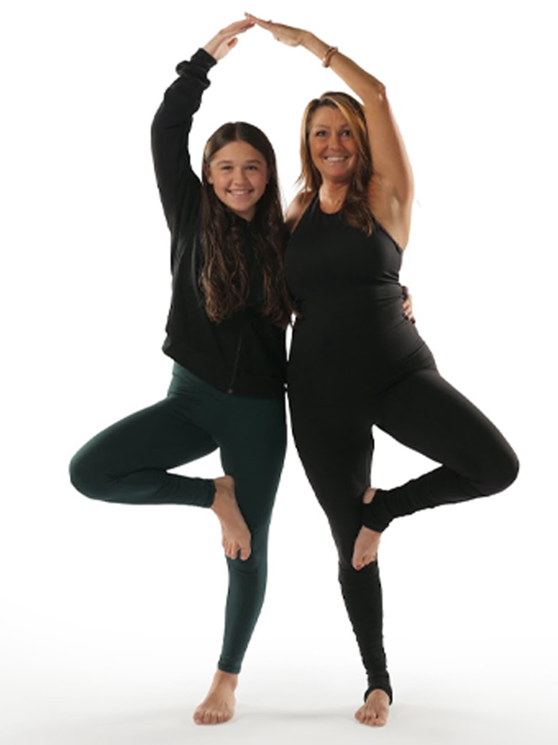 Two women practicing yoga together, standing on one leg with arms raised and touching above their heads, smiling against a white background.