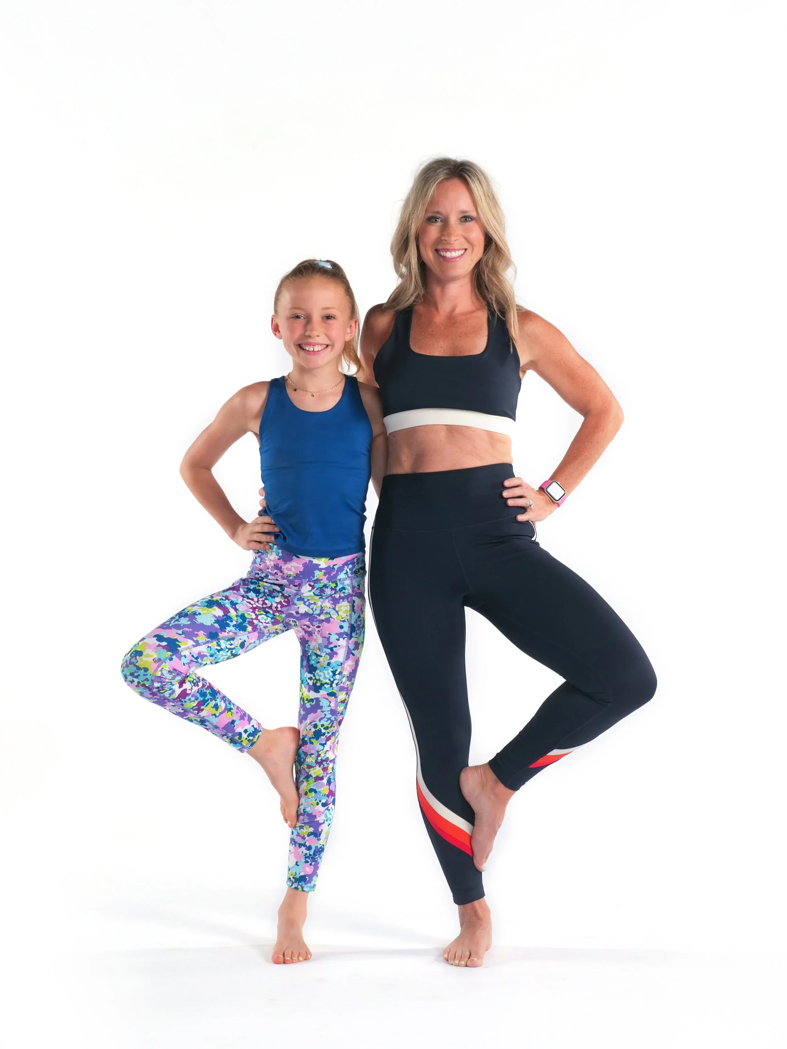 Two women practicing yoga together, one woman in colorful leggings and a blue sports top, the other in black leggings with red, white, and black stripes and a black sports bra, standing on one leg in a yoga pose against a white background.
