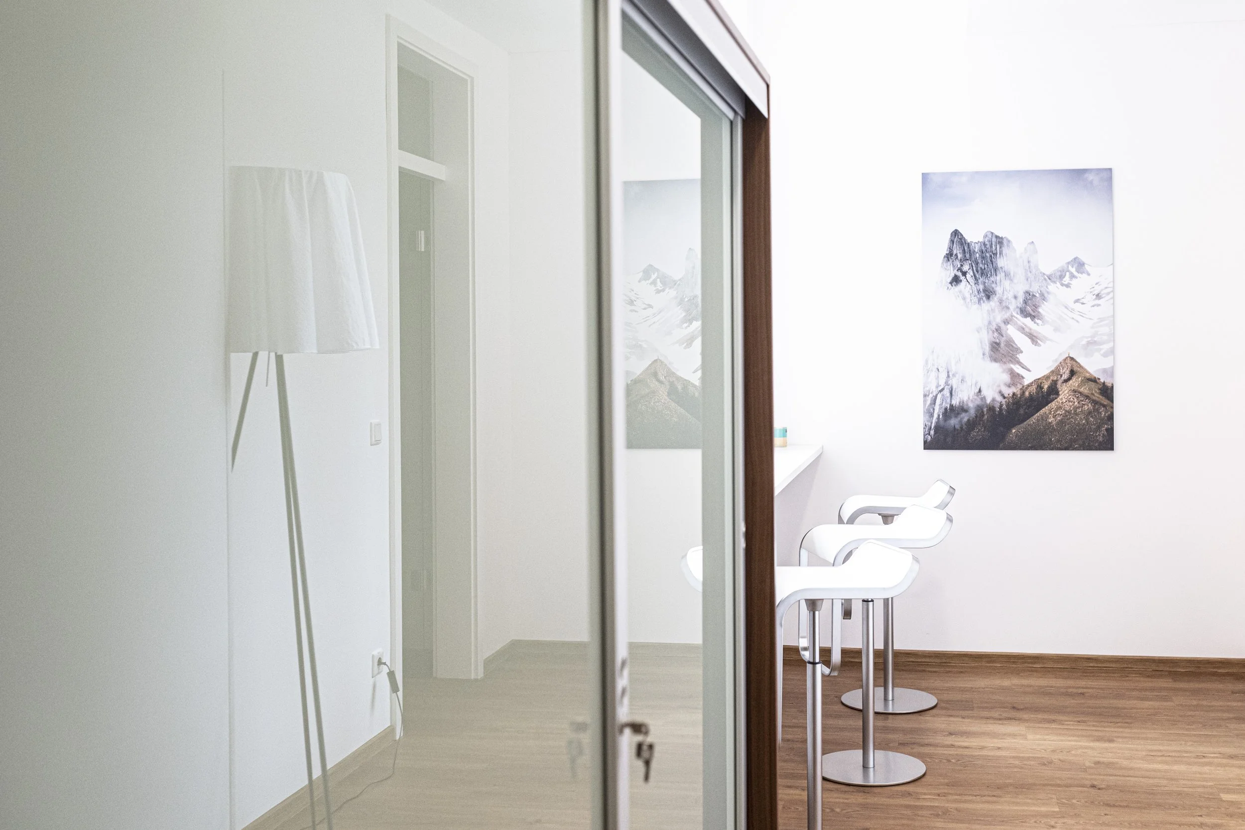 Ein heller Raum mit weißen Wänden, Holzboden, einem Wandbild von Bergen, weißen modernen Stühlen an einem hohen Bartisch, eine Stehleuchte und eine Glas-Schiebetür.