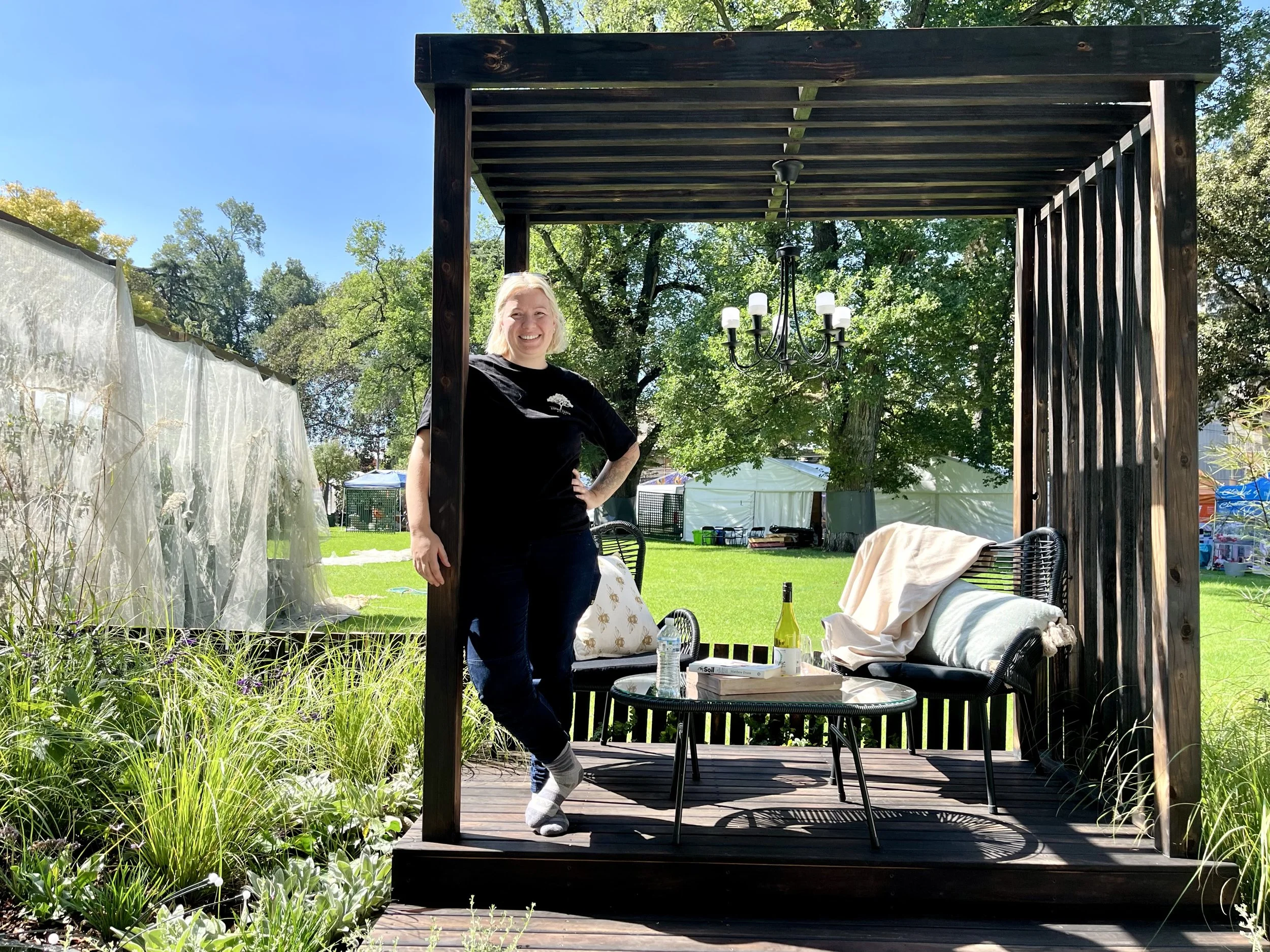 A woman standing on a wooden porch of an outdoor seating area, smiling, with a green lawn, trees, and white tents in the background. The porch has a table with a bottle of wine, a water bottle, and some food. There's a chandelier hanging overhead.