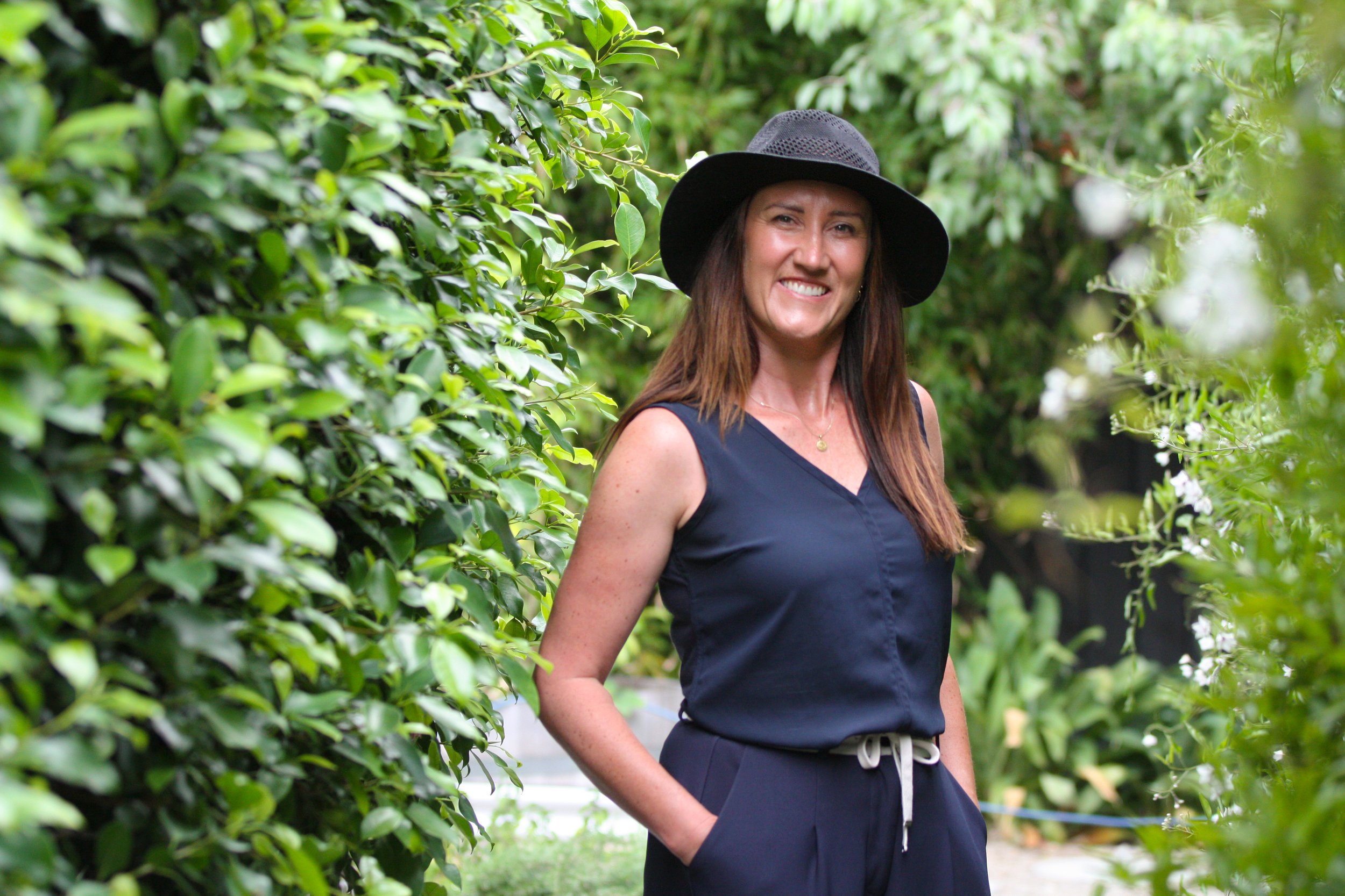 A woman standing on a wooden porch of an outdoor seating area, smiling, with a green lawn, trees, and white tents in the background. The porch has a table with a bottle of wine, a water bottle, and some food. There's a chandelier hanging overhead.