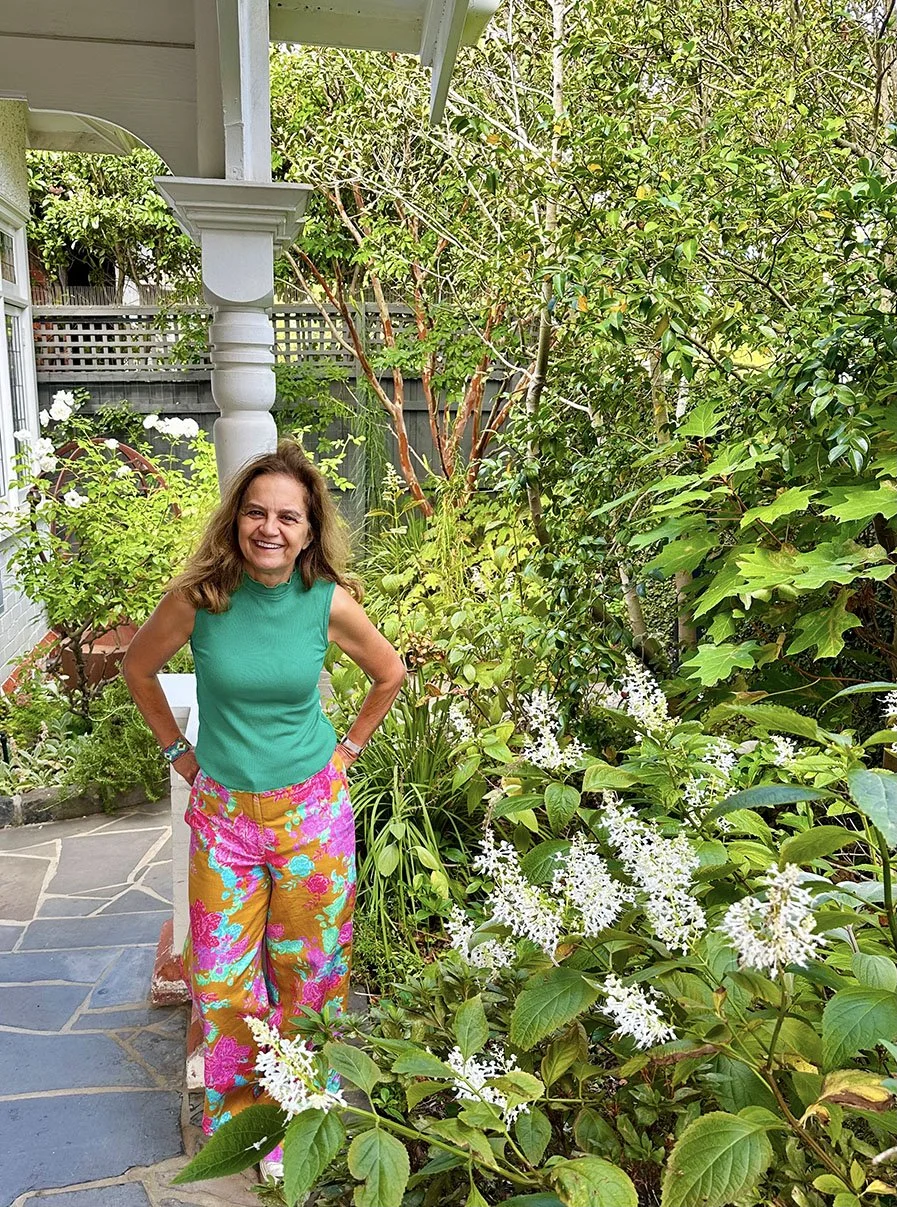 A smiling woman standing on a stone pathway in a lush garden filled with green foliage and white flowers, wearing a teal sleeveless top and colorful floral pants.