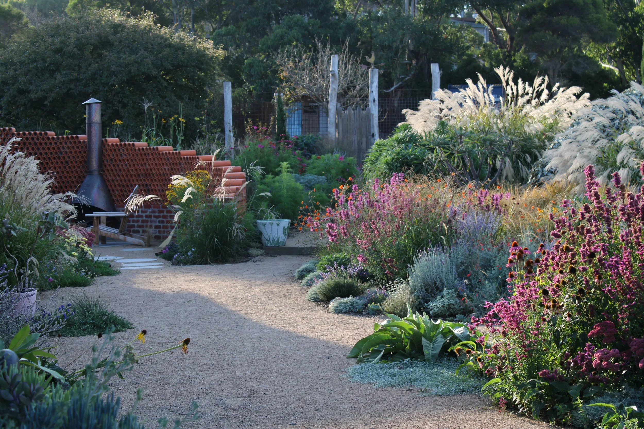 A winding dirt garden path surrounded by colorful flowering plants on both sides, with a brick and cylindrical metal stove on the left and a variety of lush green bushes and trees in the background.