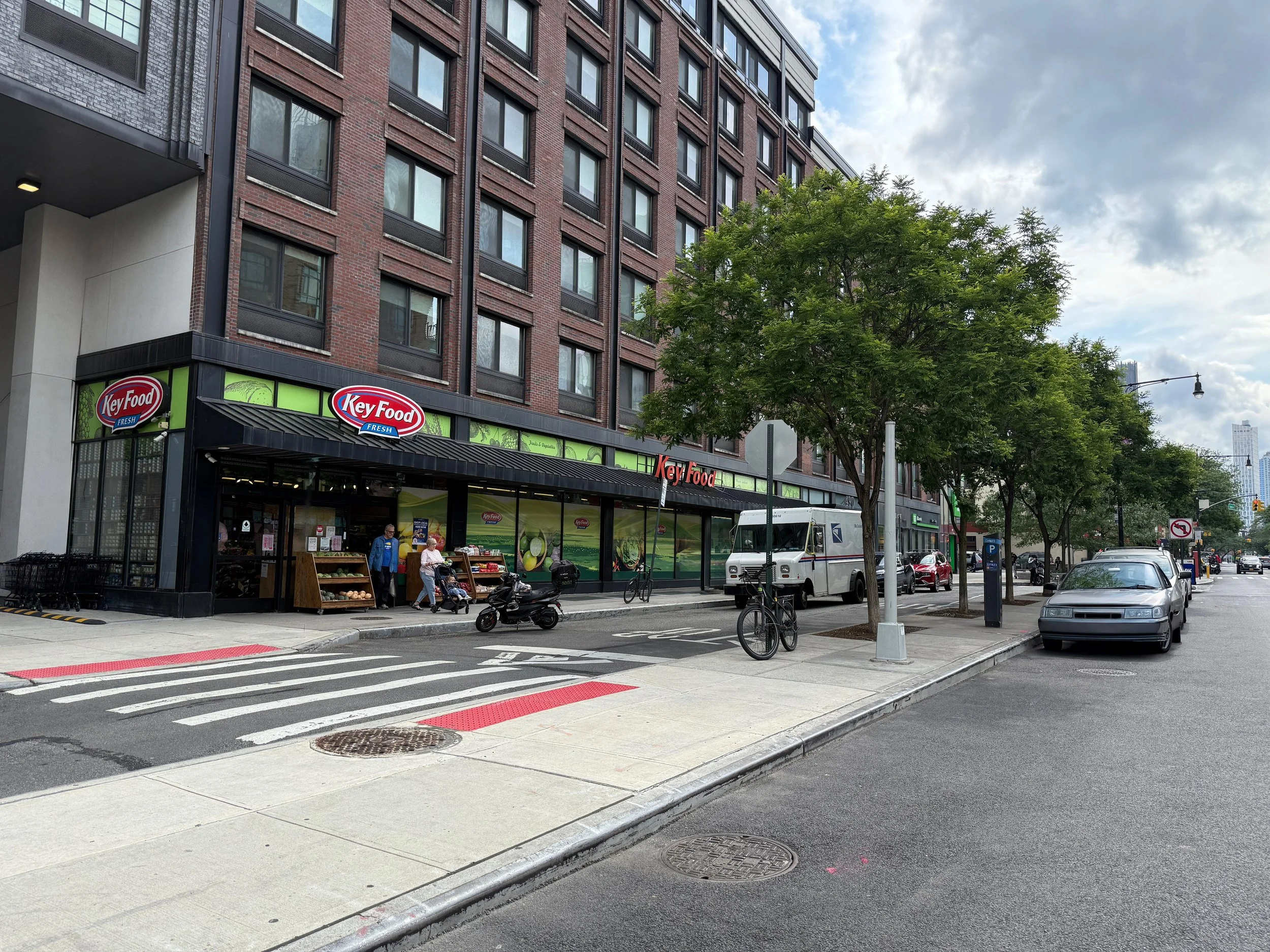 Outer view of a Key Food grocery store located on a city street, with pedestrians, parked cars, trees, and a cloudy sky.