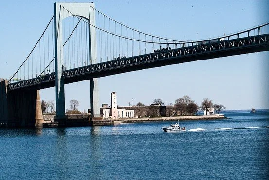 A large suspension bridge over a body of water with a boat sailing underneath and a lighthouse on the shore in the background.