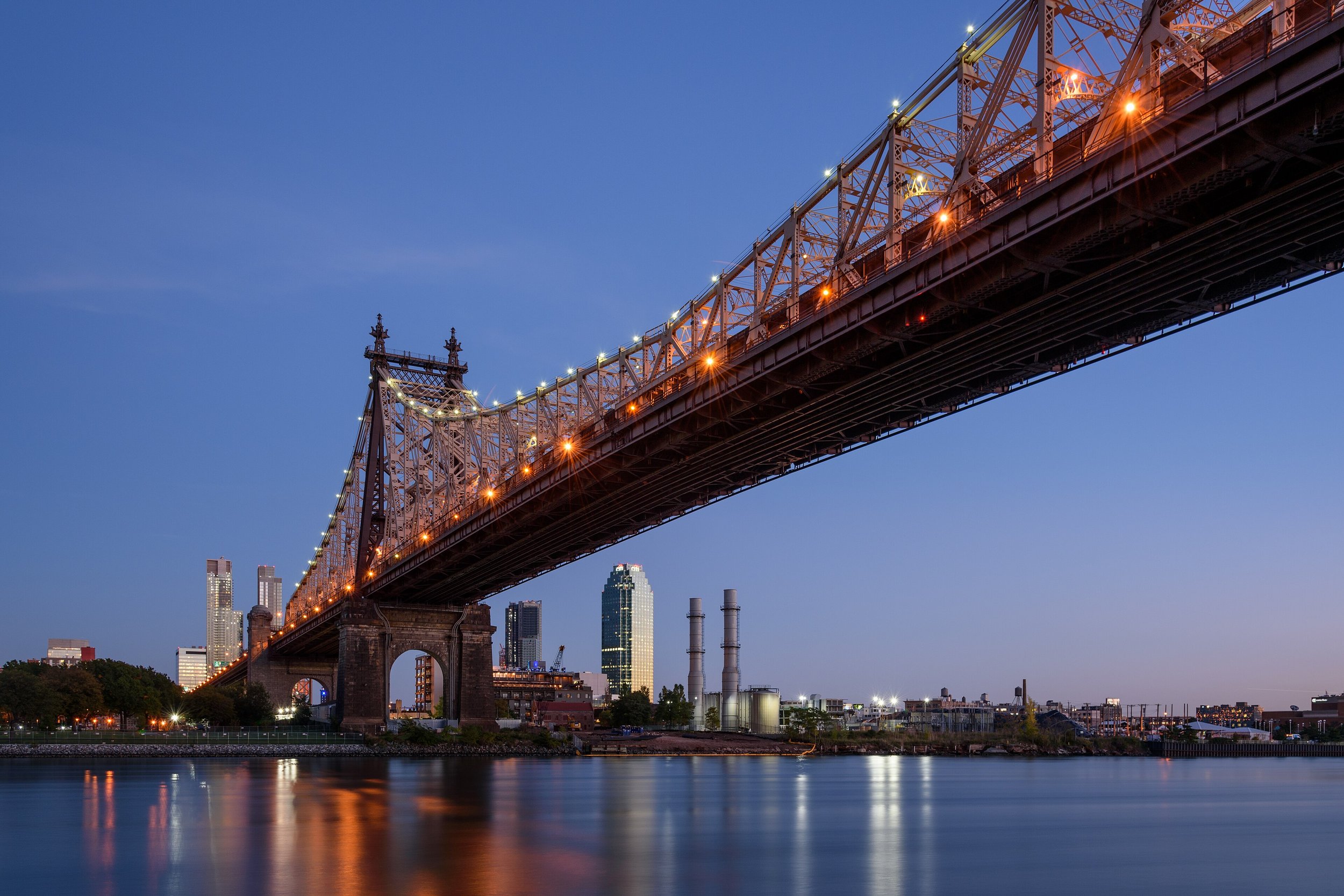 View of a bridge illuminated at dusk over a river with a city skyline in the background.