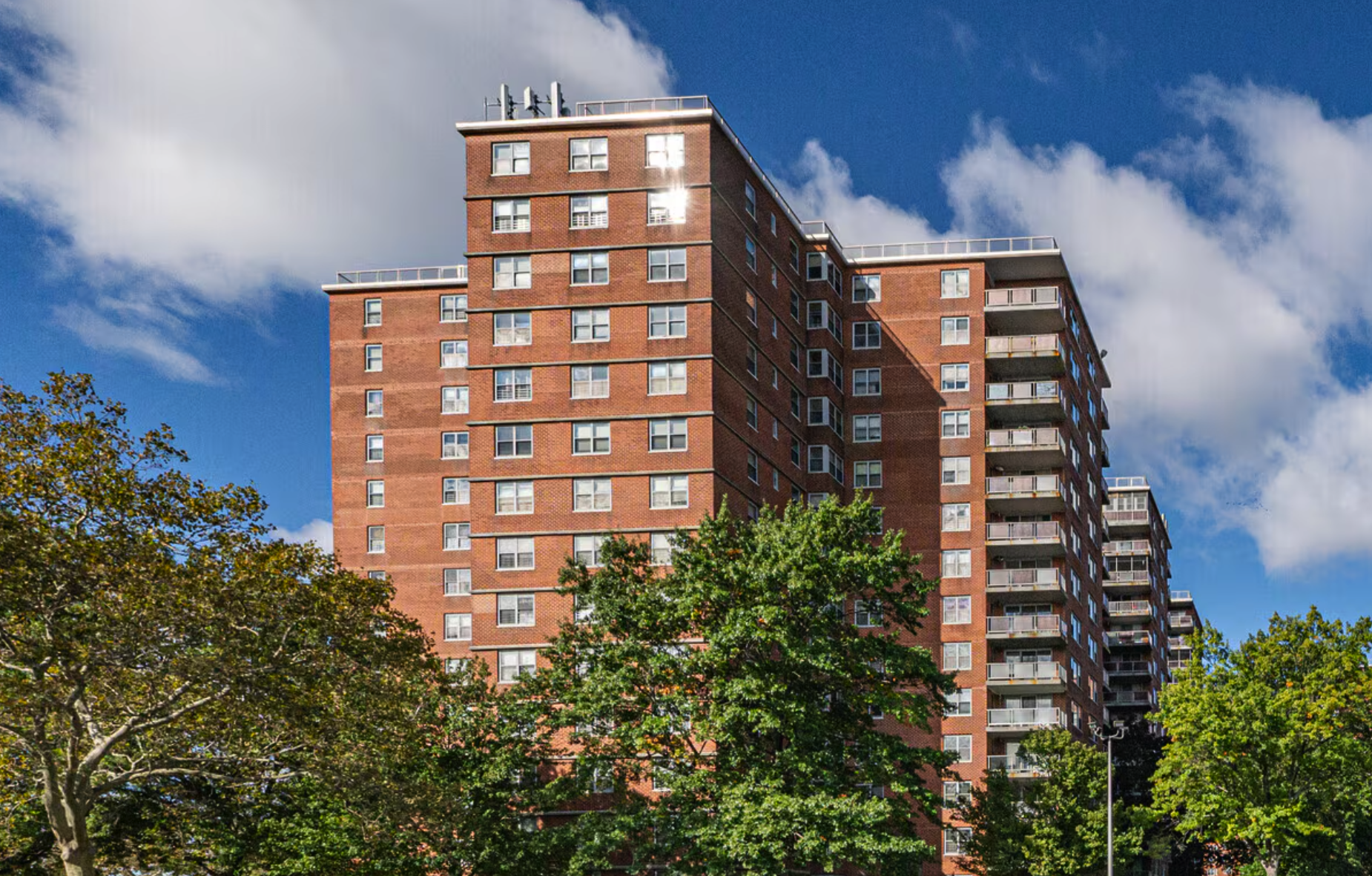 A tall red brick apartment building with multiple balconies and windows, set against a blue sky with scattered clouds, with green trees in the foreground.