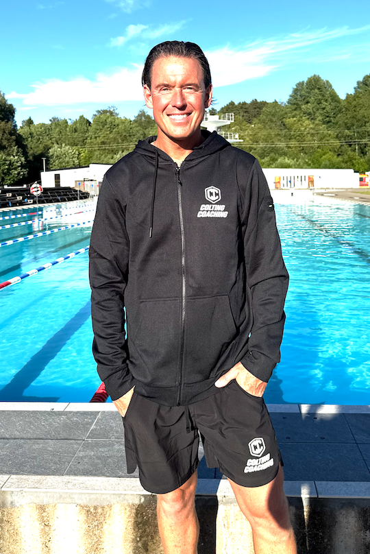 Man står vid en simhall, klädd i svart hoodie och shorts med loggor för coaching, med en utomhuspool och gröna träd i bakgrunden.