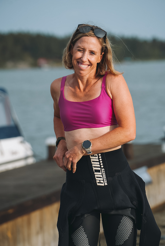 En glad kvinna i sportkläder vid vattnet, med solglasögon på huvudet och en smartwatch på handleden.