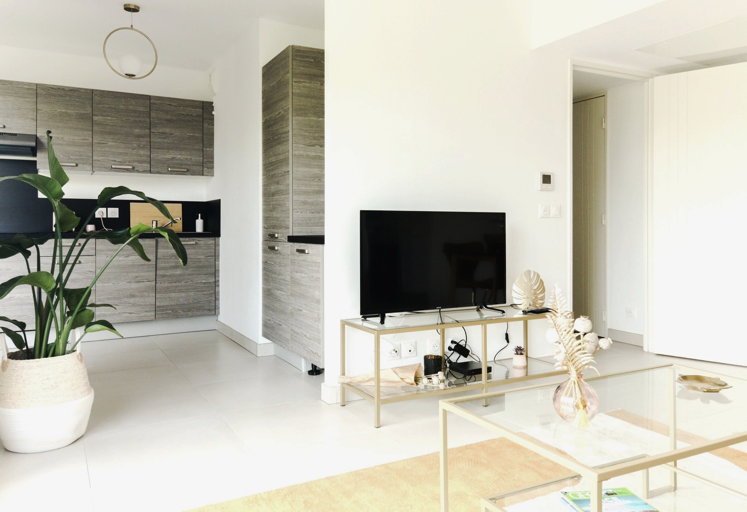 Living room with a black flat-screen TV on a glass stand, a cream-colored table with a vase of dried leaves, and a green plant in a white pot in the corner. Modern kitchen with wooden cabinets in the background.