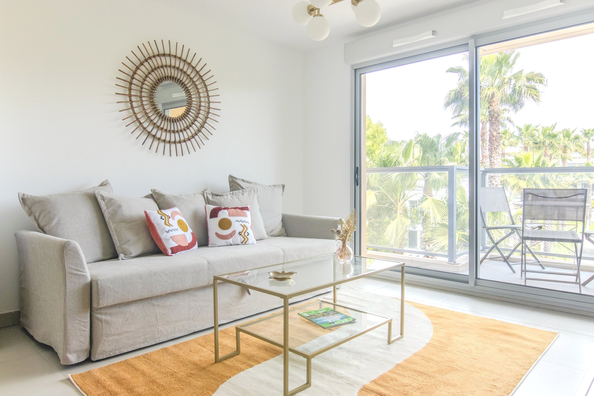 Living room with beige sofa, colorful throw pillows, glass coffee table, orange and white rug, sliding glass door leading to balcony with outdoor chairs, view of palm trees outside, modern ceiling light fixtures, and mirror on wall.