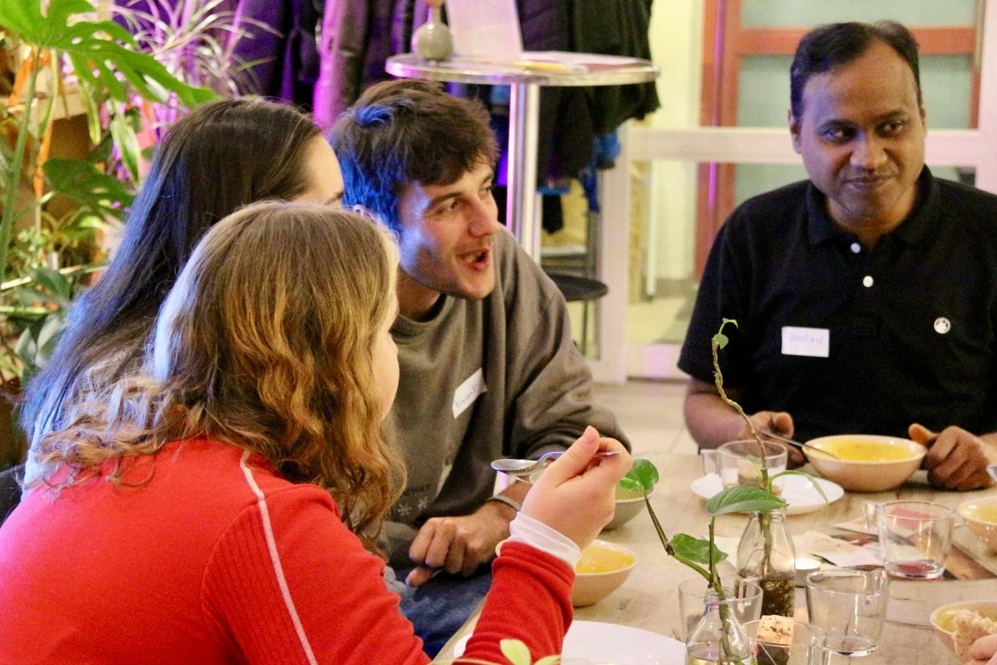 Group of four people sitting around a table, engaging in conversation with food and drinks in an indoor setting decorated with plants and colorful lighting.