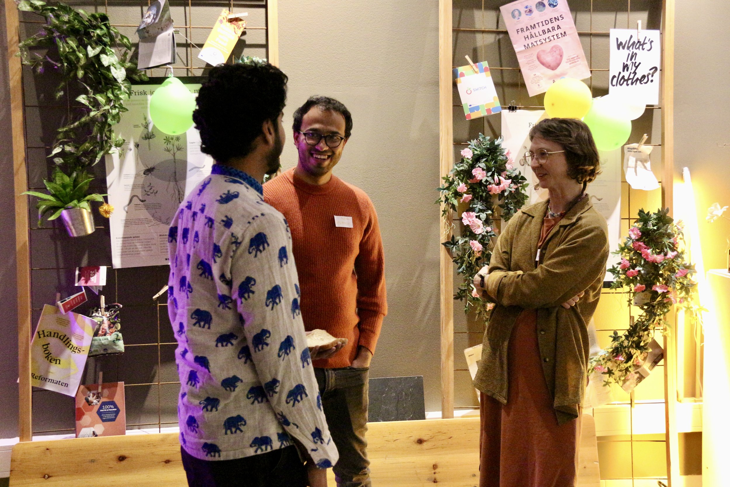 Three people standing and talking in front of a wooden display with posters, plants, balloons, and hanging notes at an indoor event.