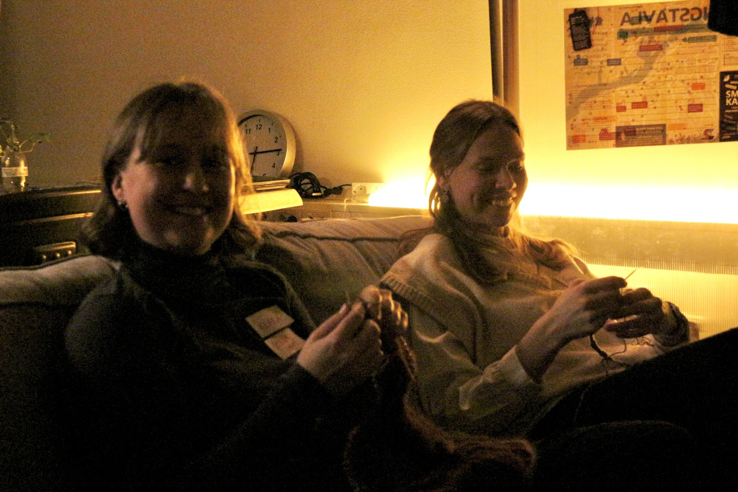 Two women sitting on a couch, smiling and holding small objects, illuminated by warm lighting, with a clock and a map on the wall behind them.