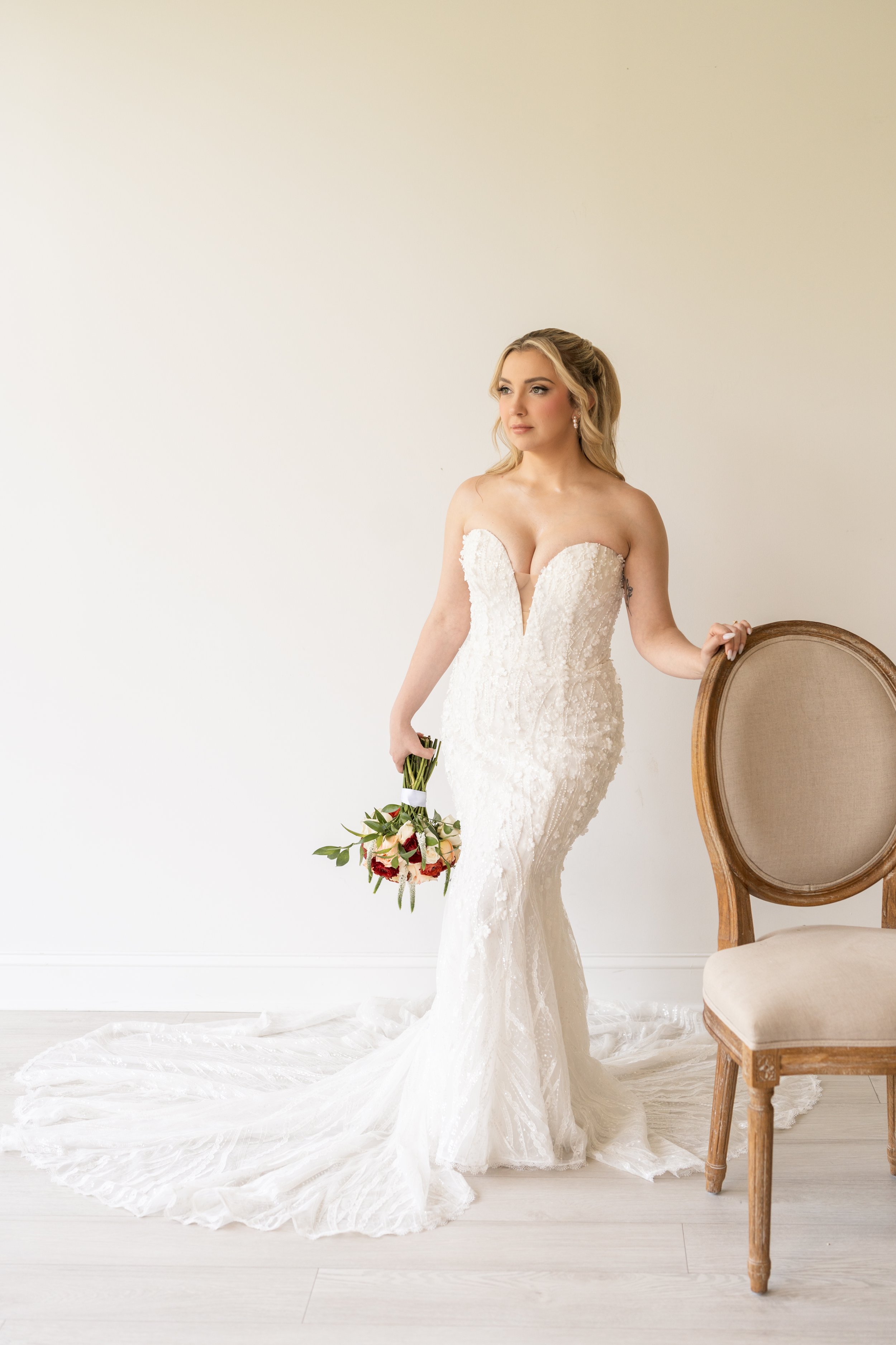 A bride in a white lace wedding gown holding a bouquet of pink and white flowers stands next to an empty vintage wooden chair against a plain white wall.