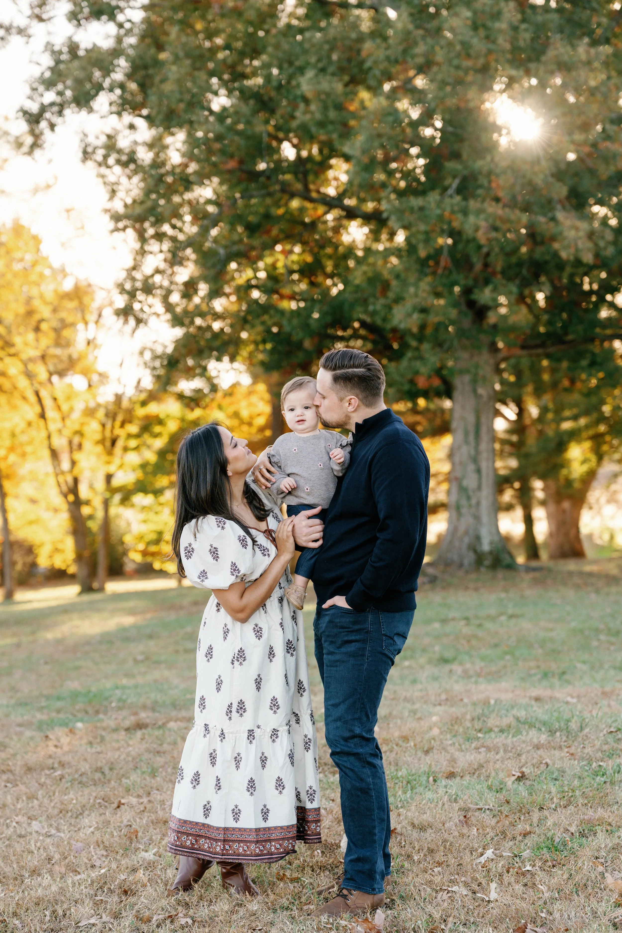 A family of three standing outdoors in a park during fall, with trees and sunlight in the background. The woman is holding a young girl, and the man is kissing the girl's cheek.