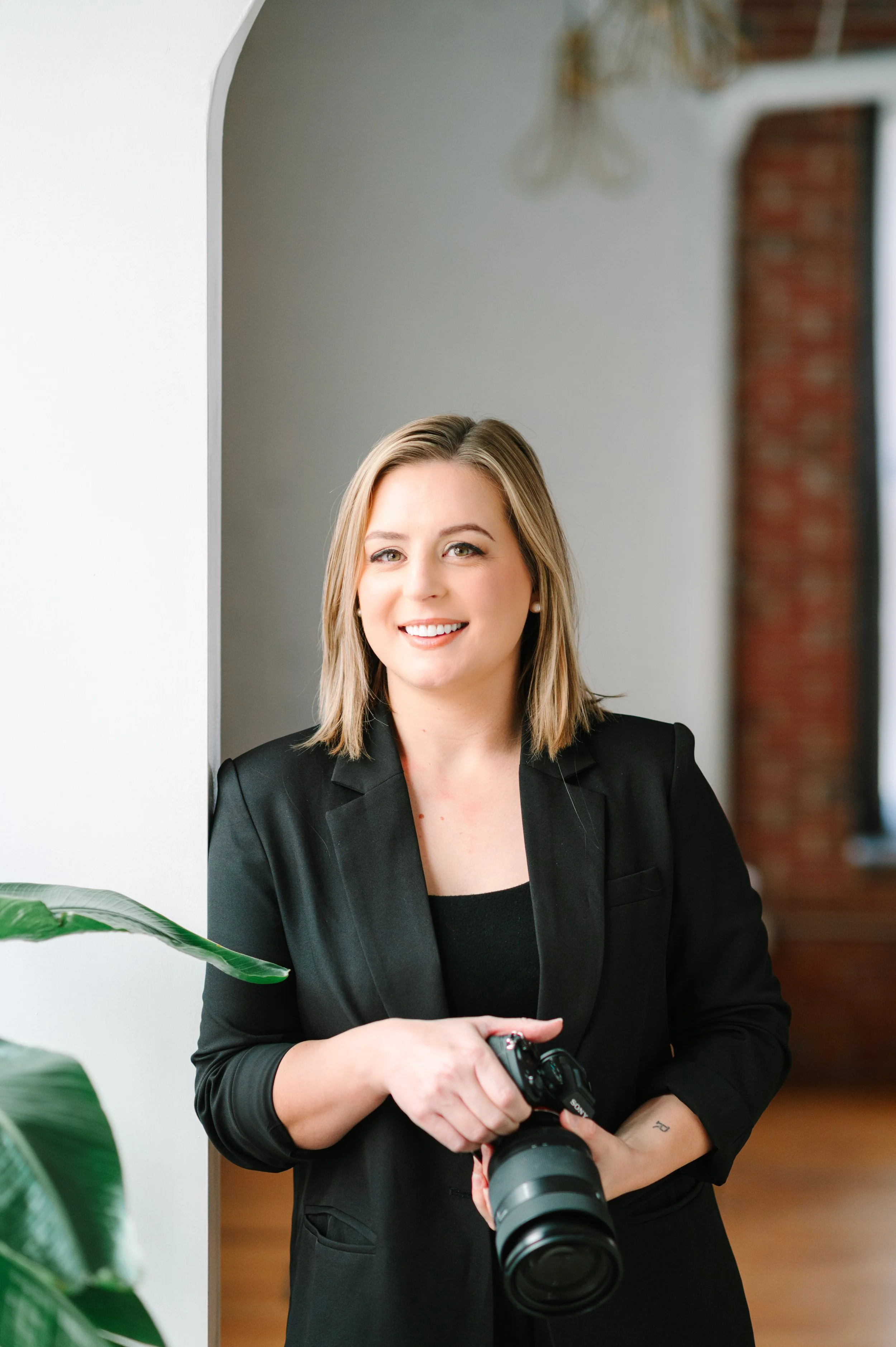 A woman with shoulder-length blonde hair in a black blazer holding a camera, smiling, standing next to a white wall with large green leaves in front.