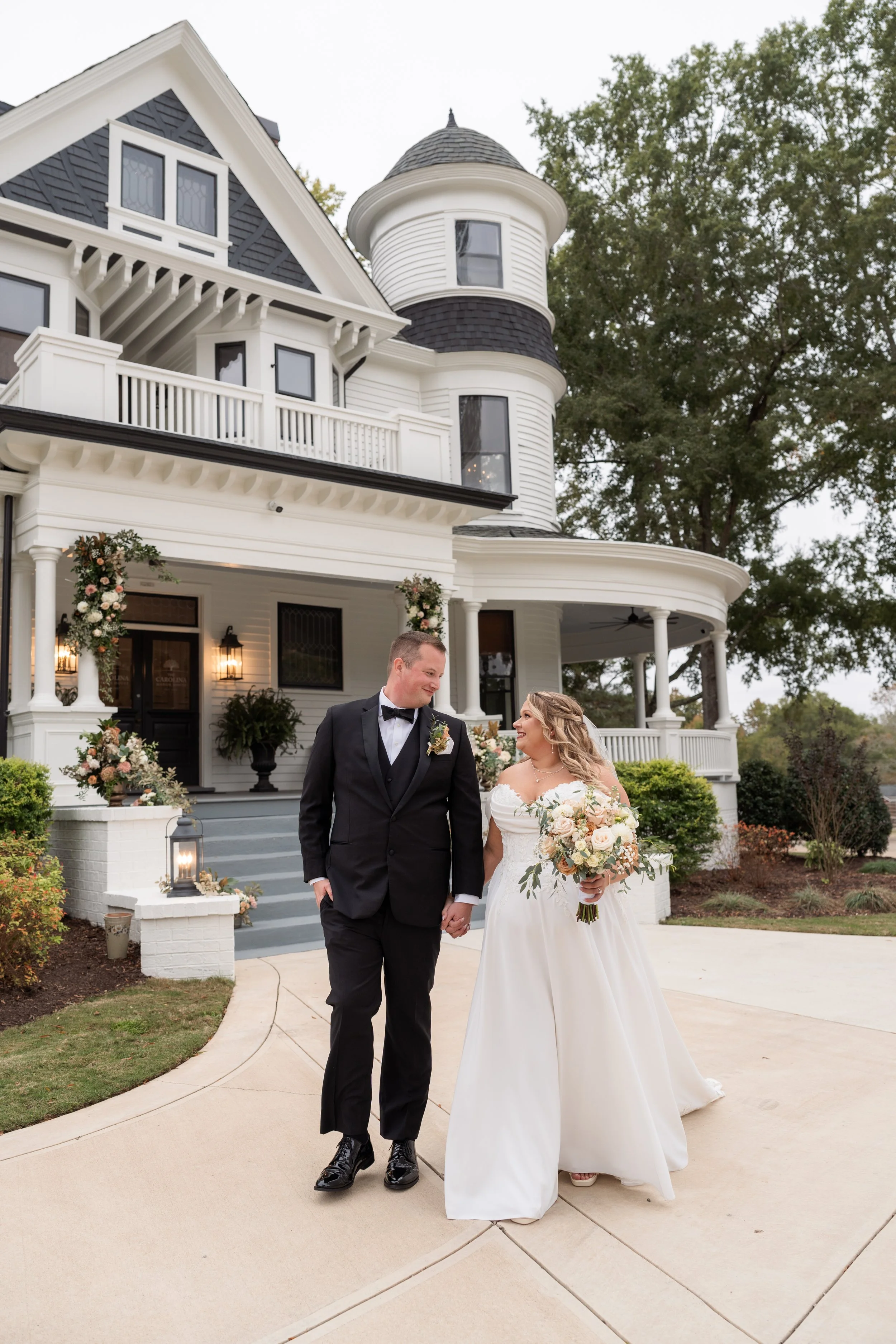 A bride and groom walk hand-in-hand outside a Victorian-style white house decorated for a wedding, holding a floral bouquet.