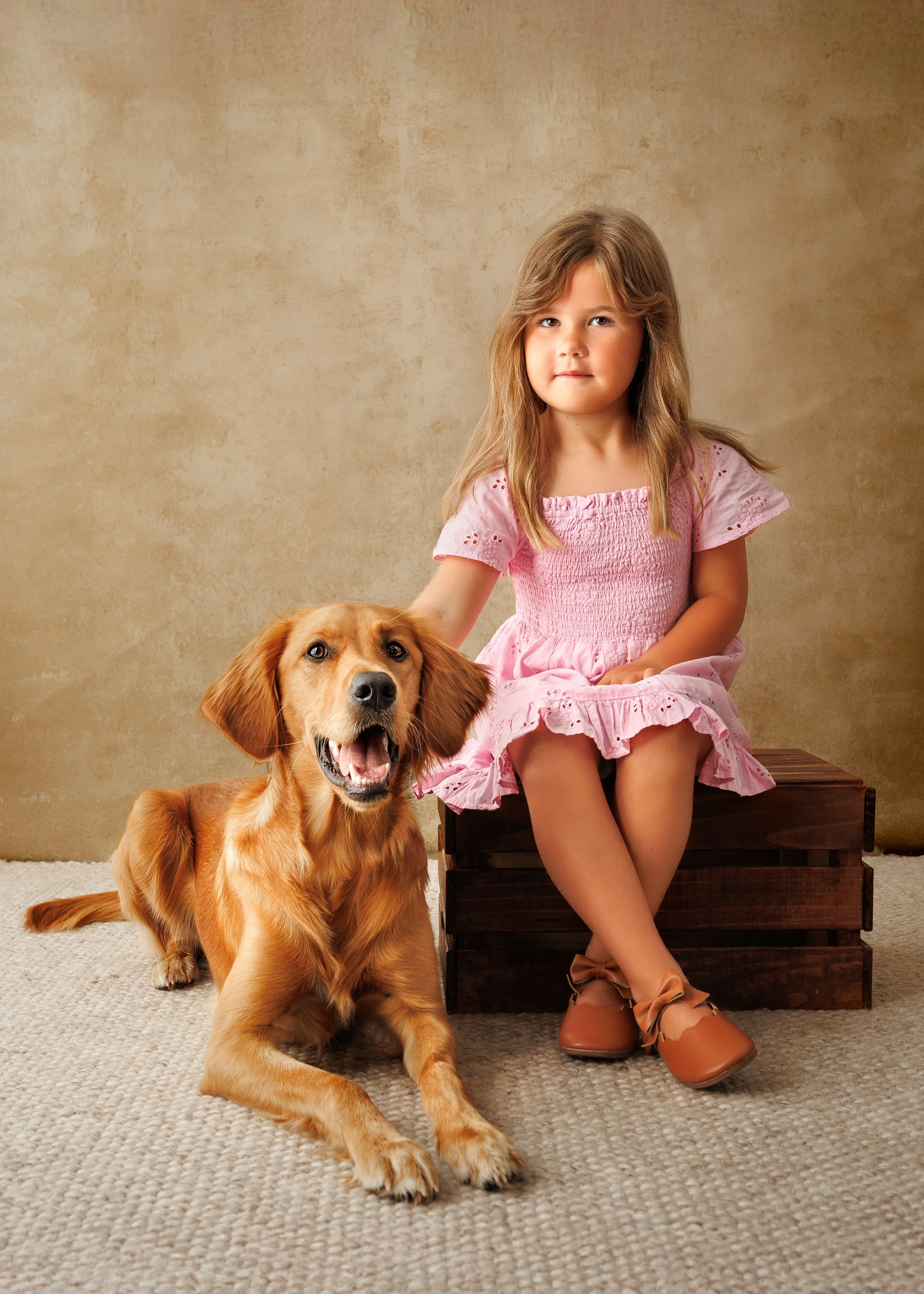 A young girl with long light brown hair wearing a pink dress sitting on a wooden box, petting a happy golden retriever laying on a beige carpeted floor against a textured beige background photographed by Margaret River and Busselton dog photographer 