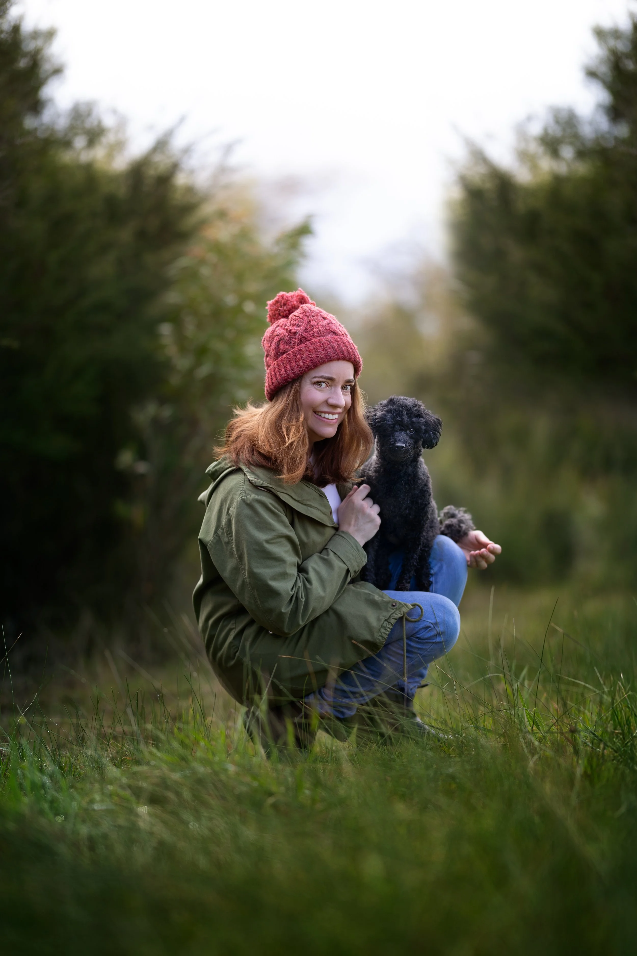 A woman with red hair, wearing a red knit hat and green jacket, smiling while squatting outdoors next to a small black dog in a grassy area surrounded by trees in Margaret River