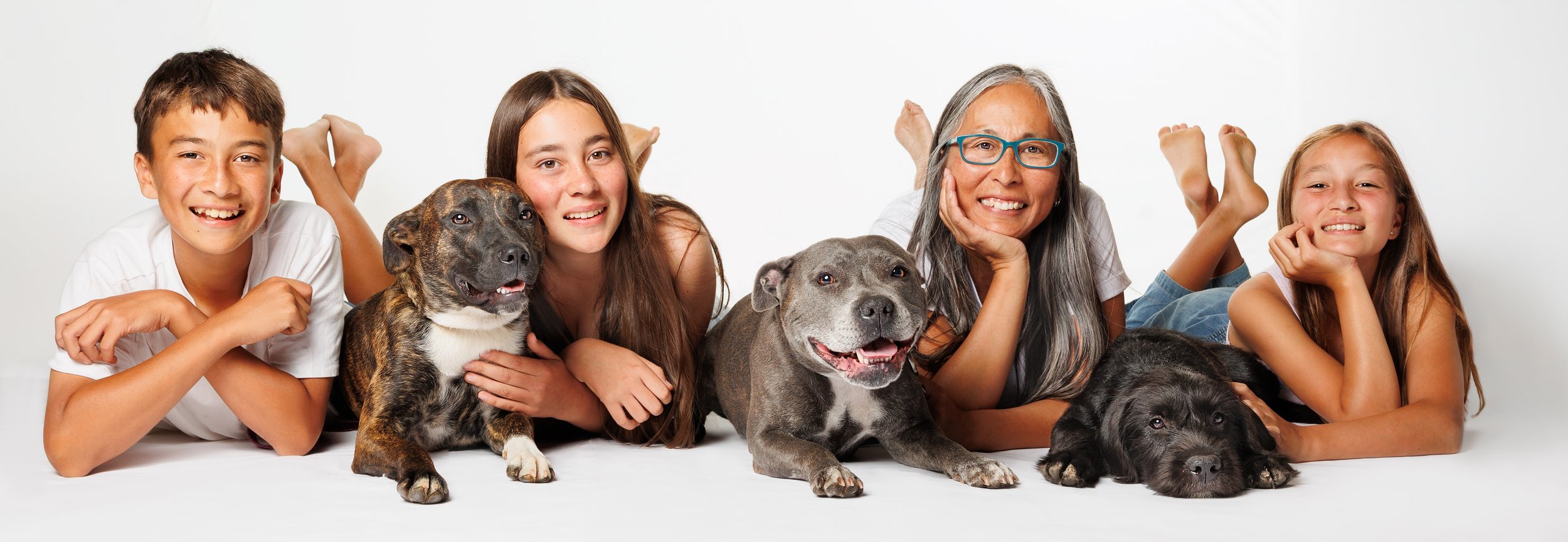 Family of three children, an elderly woman, and three dogs lying on the floor against a plain white background.