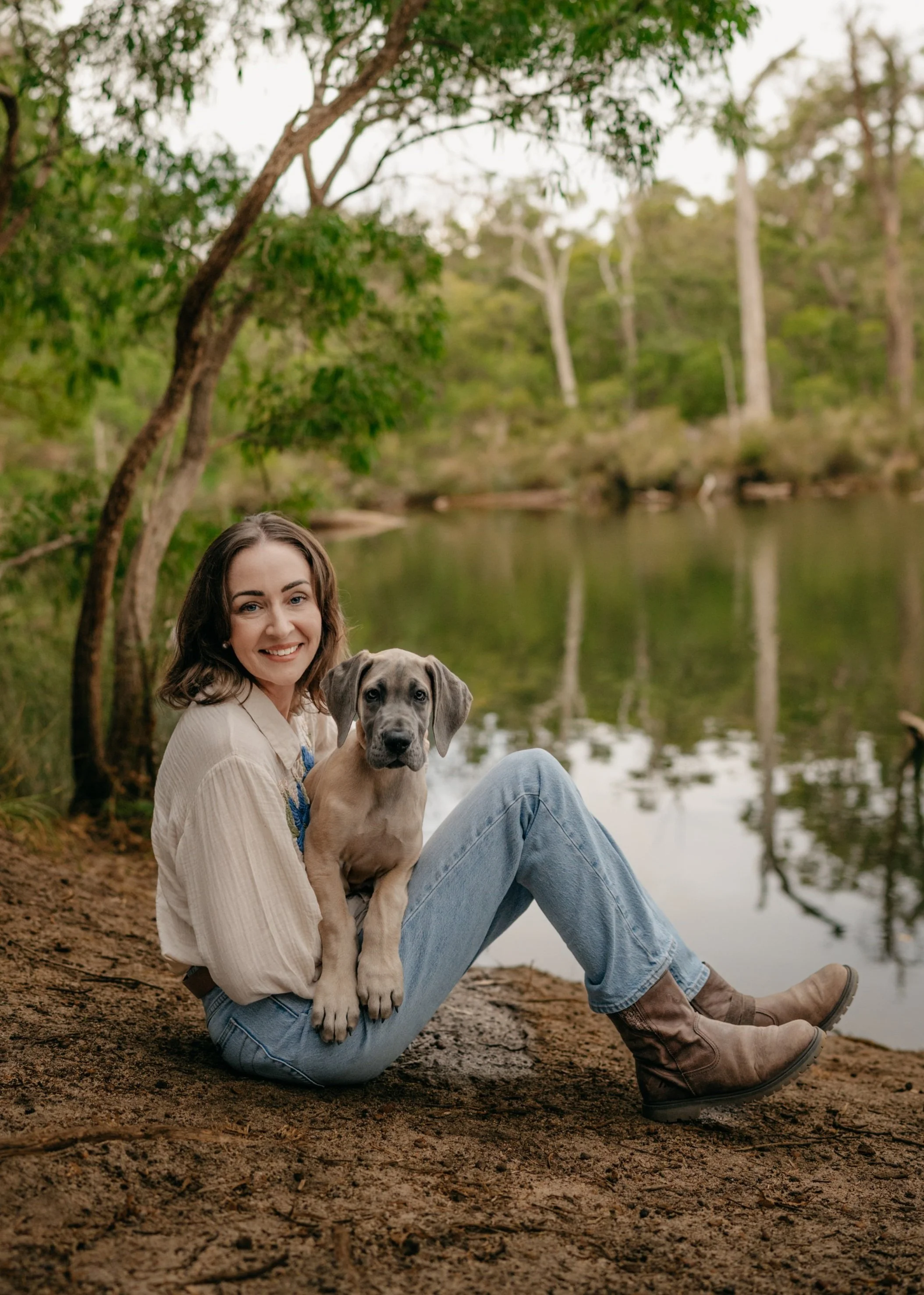 A woman sitting on the ground by a lake, holding a puppy in her lap, surrounded by trees and nature.