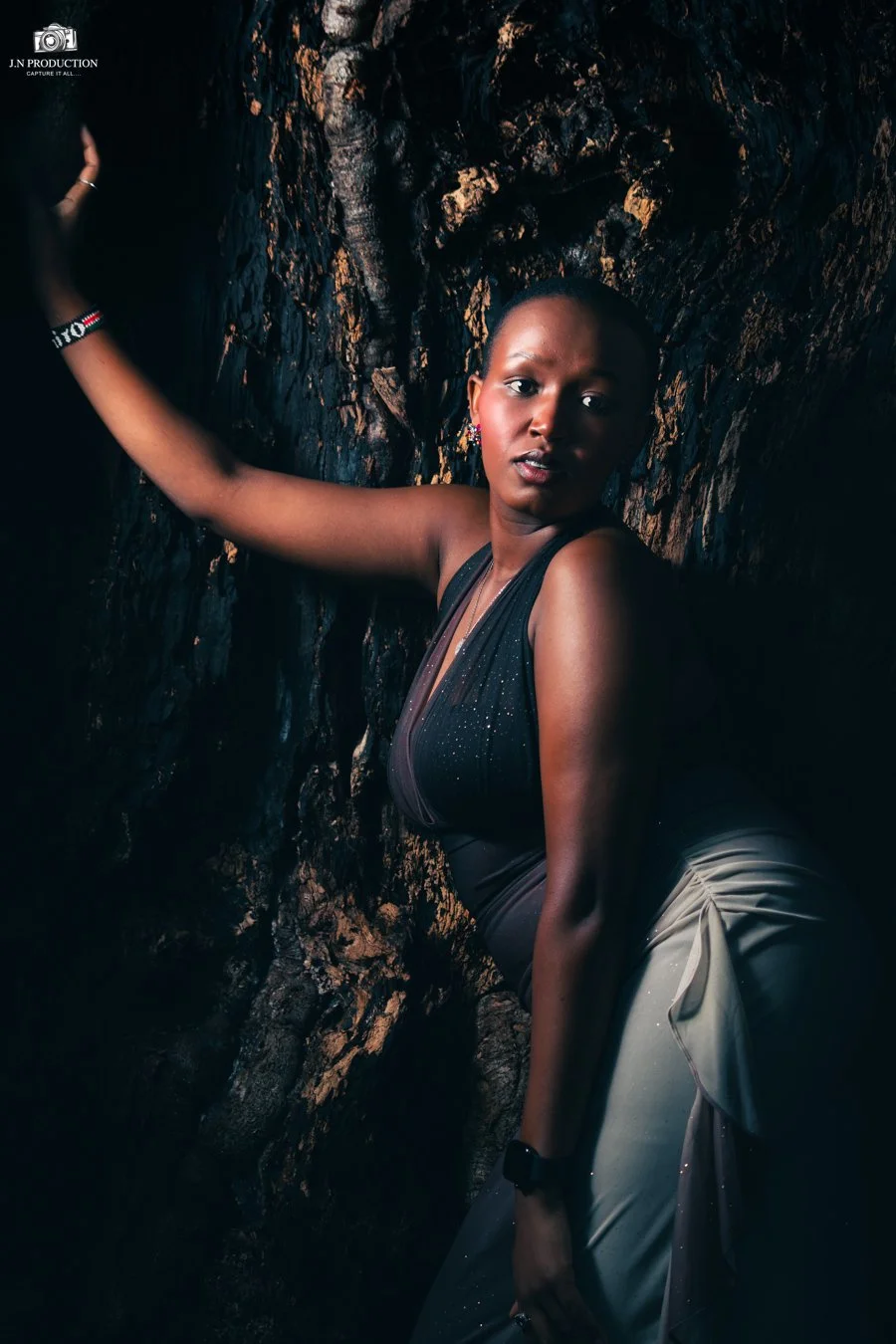 A woman wearing a black top and light-colored skirt leans against a dark, textured rock wall, looking directly at the camera.