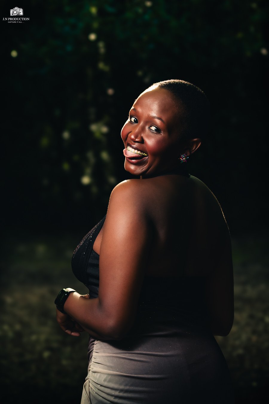 A smiling woman with a short haircut, wearing earrings and a sleeveless top, outdoors at night with a dark, leafy background.