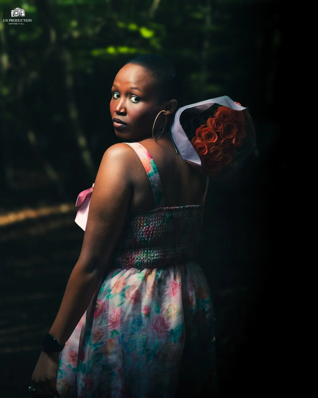 A woman with dark skin and short hair holding a bouquet of red roses on her shoulder, wearing a colorful floral dress, in a dimly lit outdoor setting.
