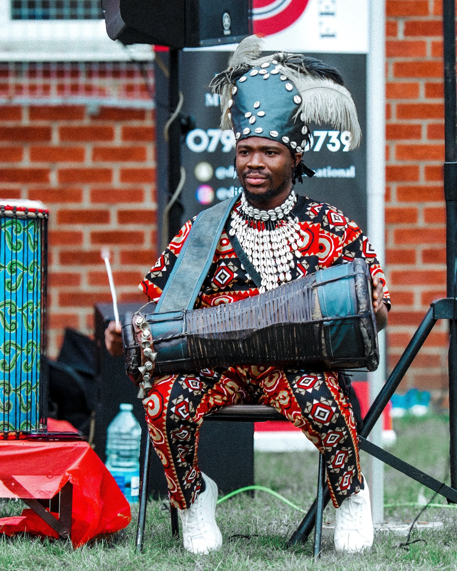 A man wearing traditional African attire, including a colorful patterned shirt, pants, and a large headpiece with shells, is seated and playing a traditional string instrument.