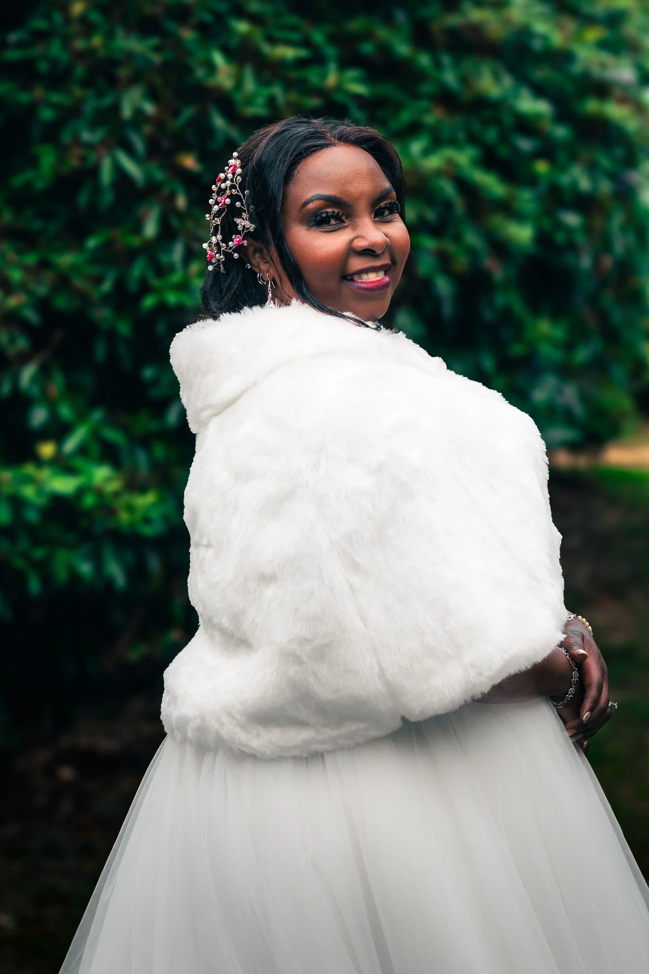 A woman dressed in a white tulle gown with a white faux fur shawl, standing outdoors in front of green foliage, smiling at the camera.