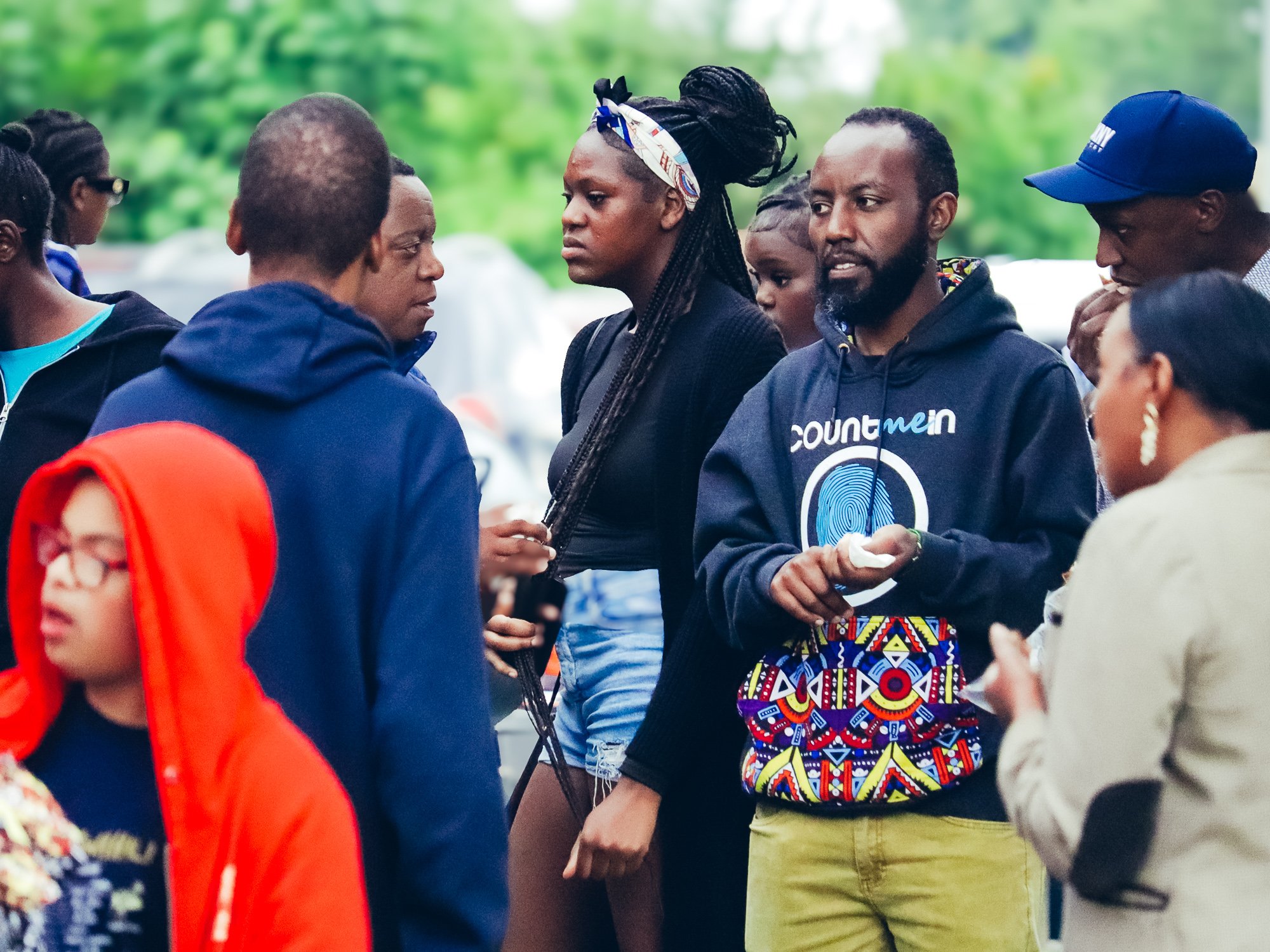 A group of people gathered outdoors, engaging in conversation, including a man in a black hoodie with a 