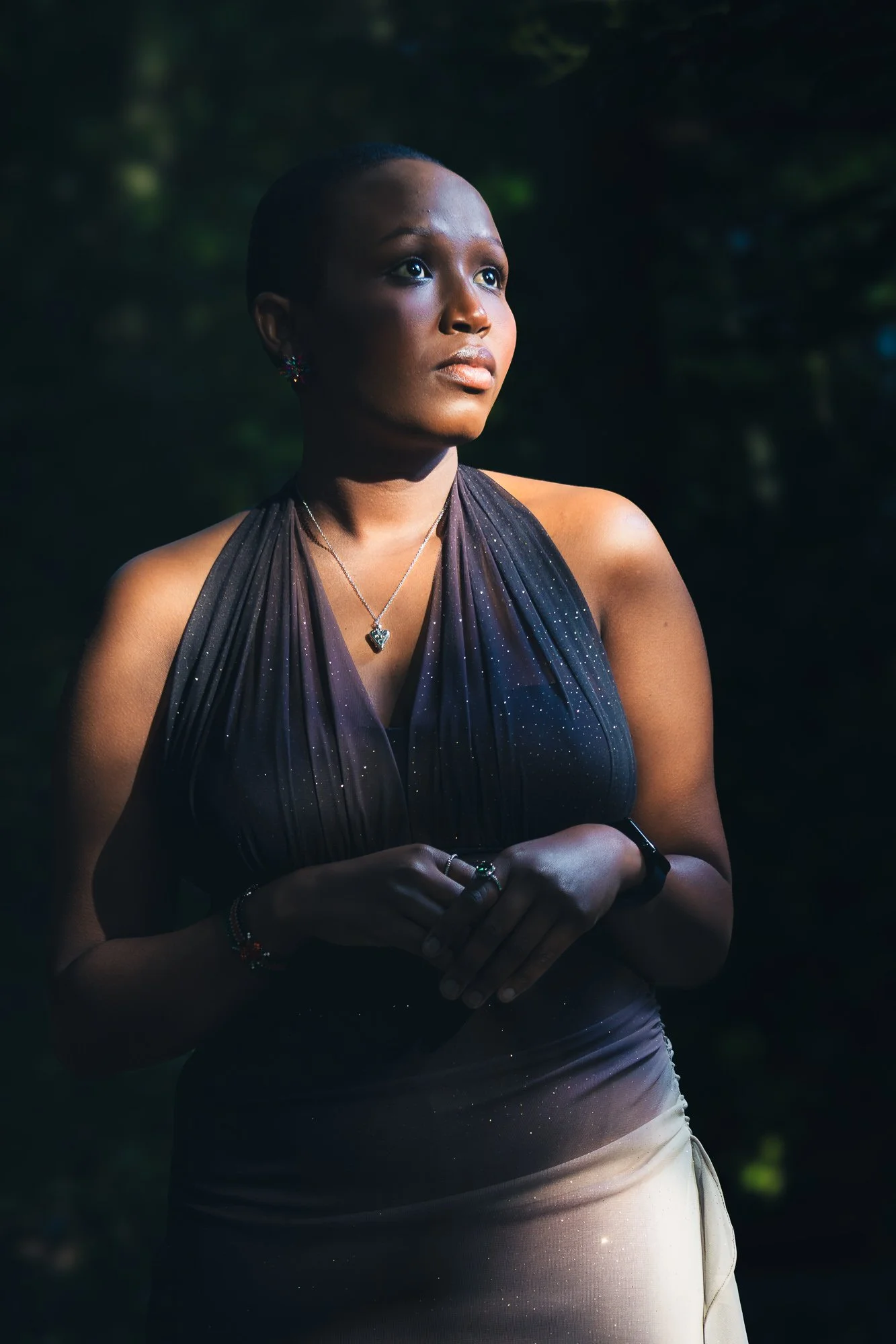 A young woman standing outdoors with dark background, looking off to the side, wearing a sleeveless black dress with sparkles, jewelry, and a necklace with a heart-shaped pendant.