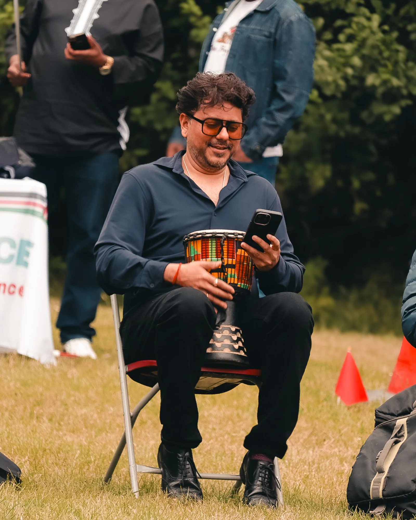 A man sitting on a chair outdoors, playing a small drum and looking at his phone, with two other people standing nearby in a grassy area.