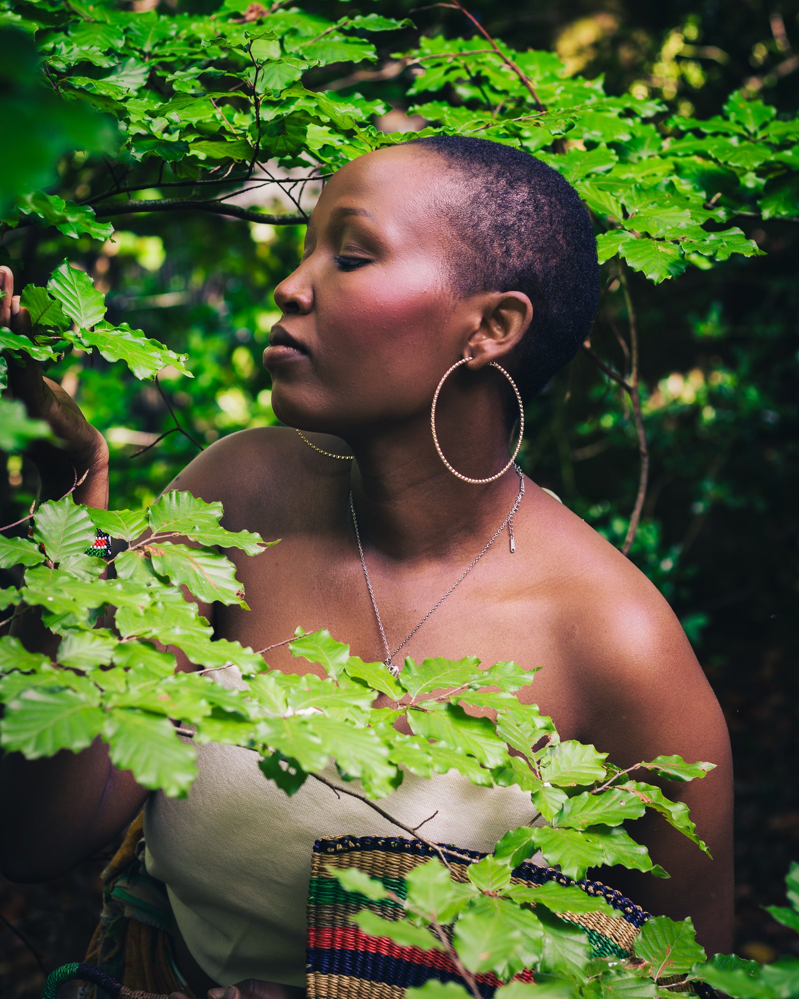 A woman with a short, natural haircut and large hoop earrings stands among green leaves in a lush outdoor setting, with closed eyes and a serene expression.