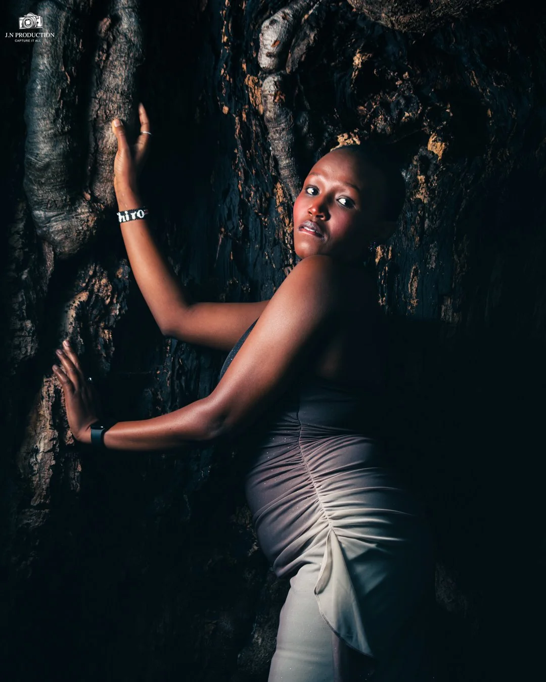 A woman with dark skin and short hair leans against a textured dark rock wall, looking at the camera with a serious expression. She is wearing a sleeveless, ruched beige dress and a beaded bracelet.