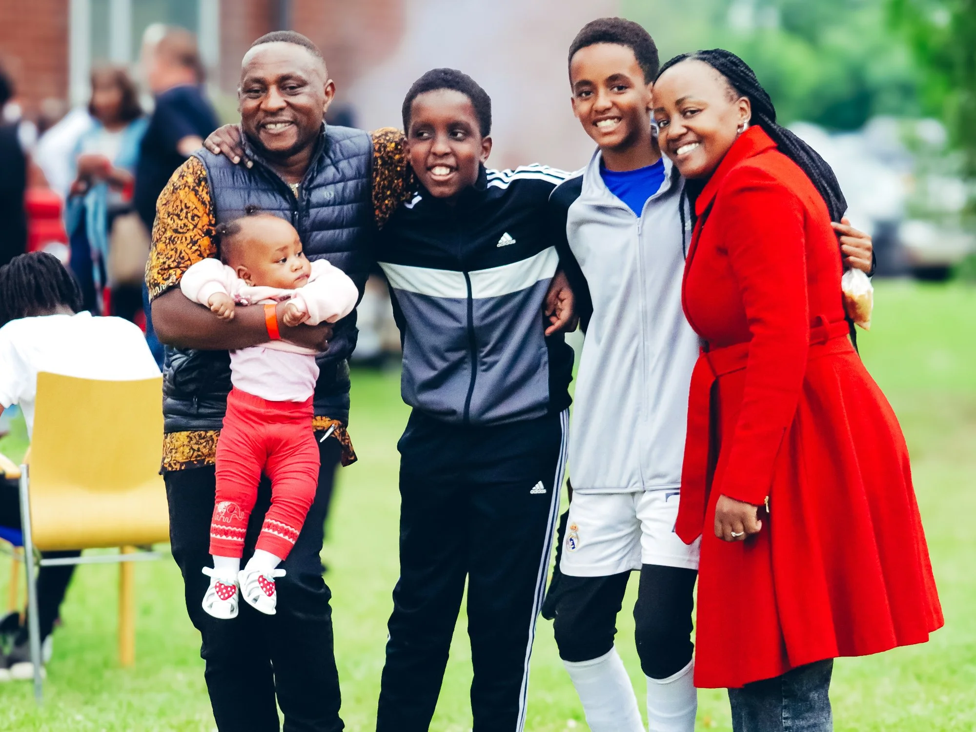Family of six standing outdoors, smiling, with green grass and trees in the background. One woman in a red dress, a man, three boys, and a baby girl.