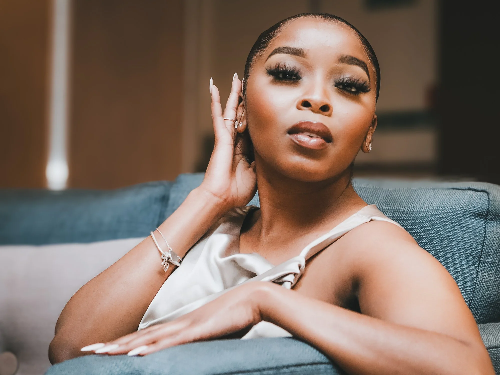 A young woman with dark hair styled in a sleek bun, wearing makeup, a sleeveless satin top, and jewelry, sitting on a blue couch in an indoor setting, posing with her hand touching her face and looking at the camera.